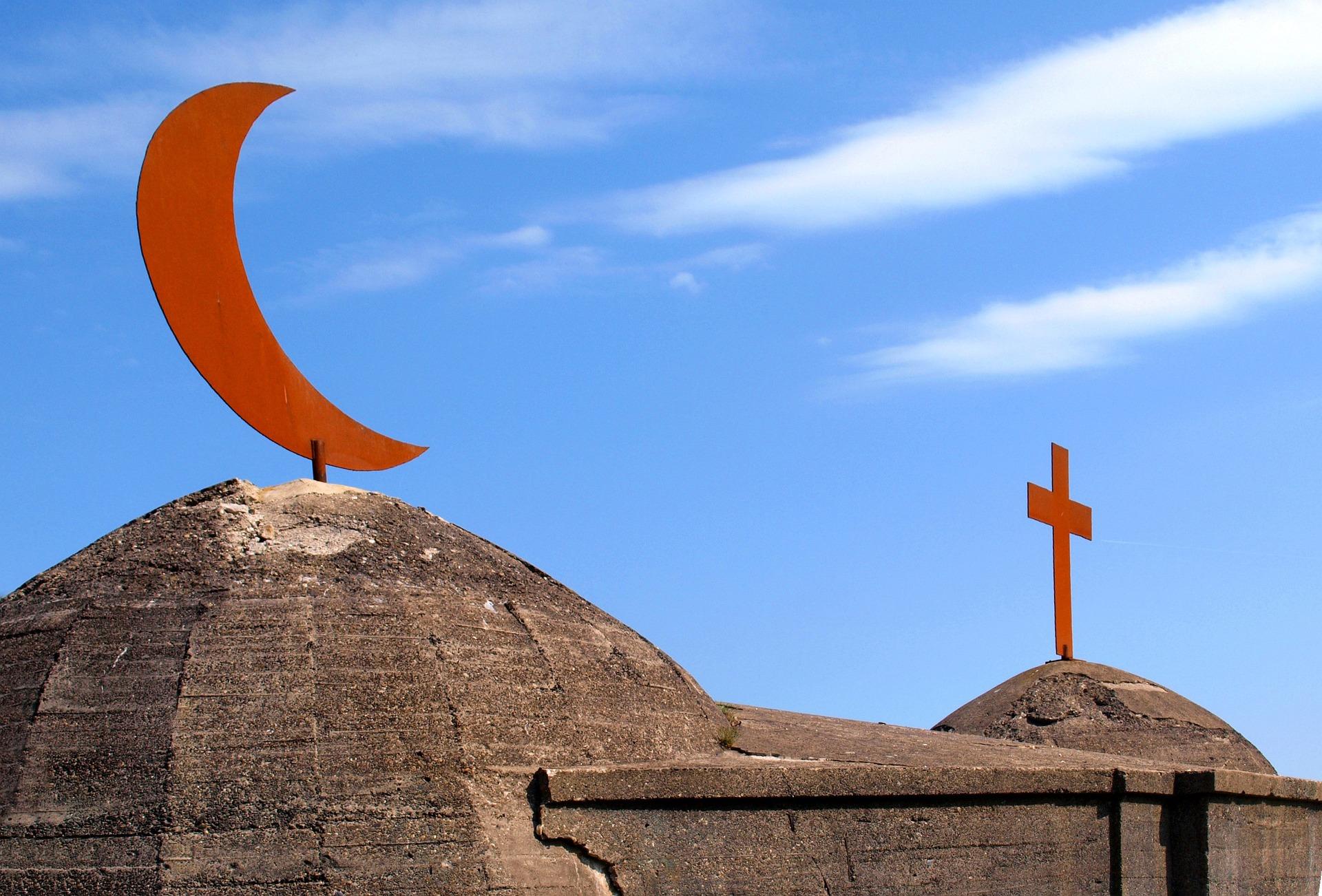Zwei Dächer sind zu sehen: Vorne eine Kuppel mit einem Halbmond, hinten eine Kuppel mit einem Kreuz. Darüber blauer Himmel.