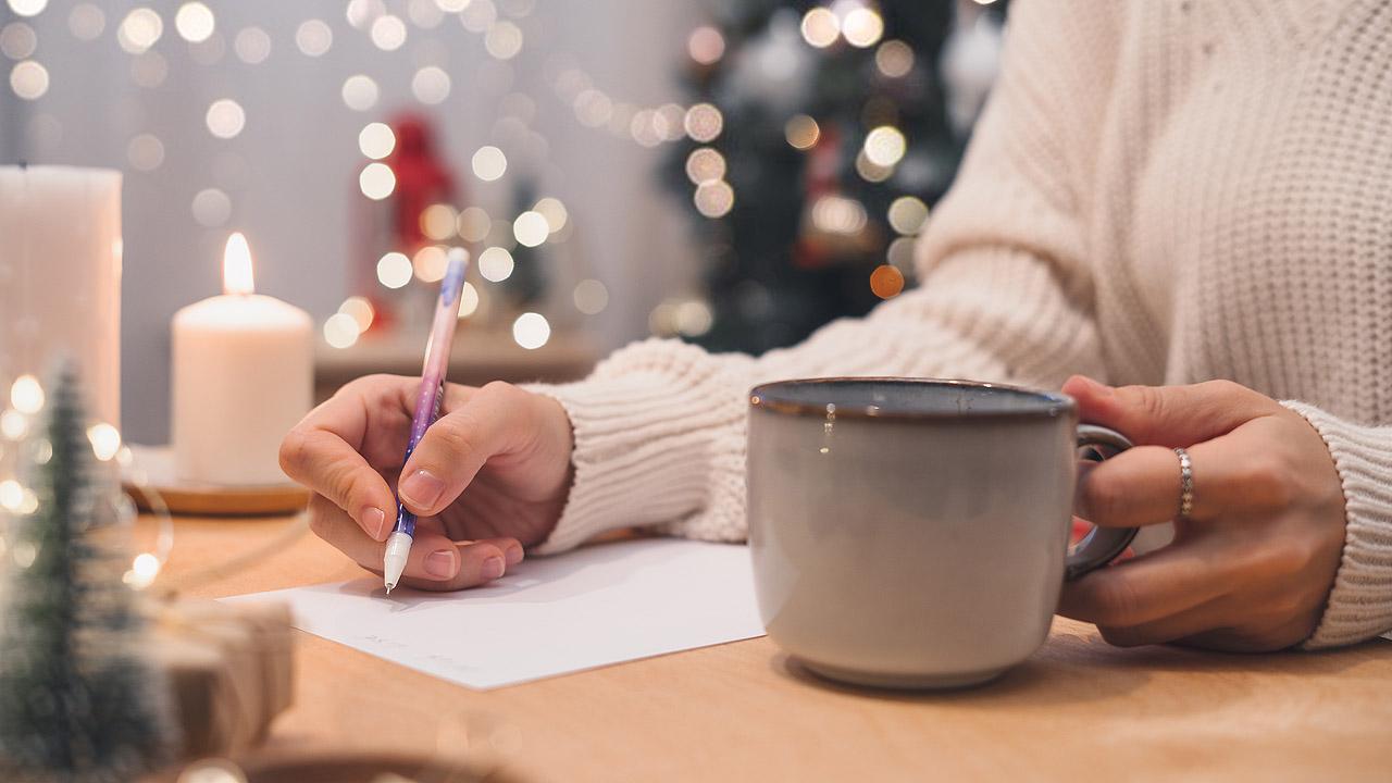 Hand einer Frau die schreibt und eine große Tasse mit einem heißen Getränk. Dahinter Lichter und Kerzen, die noch auf Weihnachten hindeuten