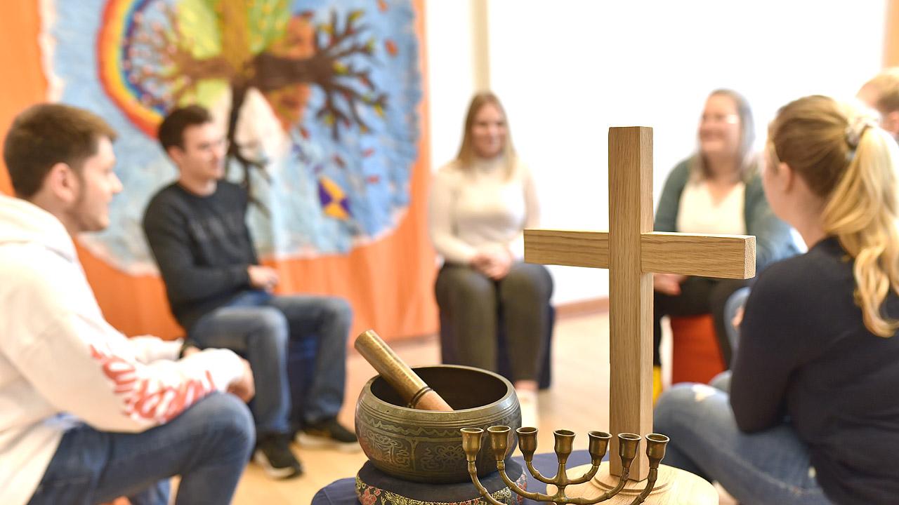 Meditation Oberstufenschüler sitzen im Kreis, davor sind ein christliches Kreuz, eine Klangschale und ein siebenarmiger Leuchter