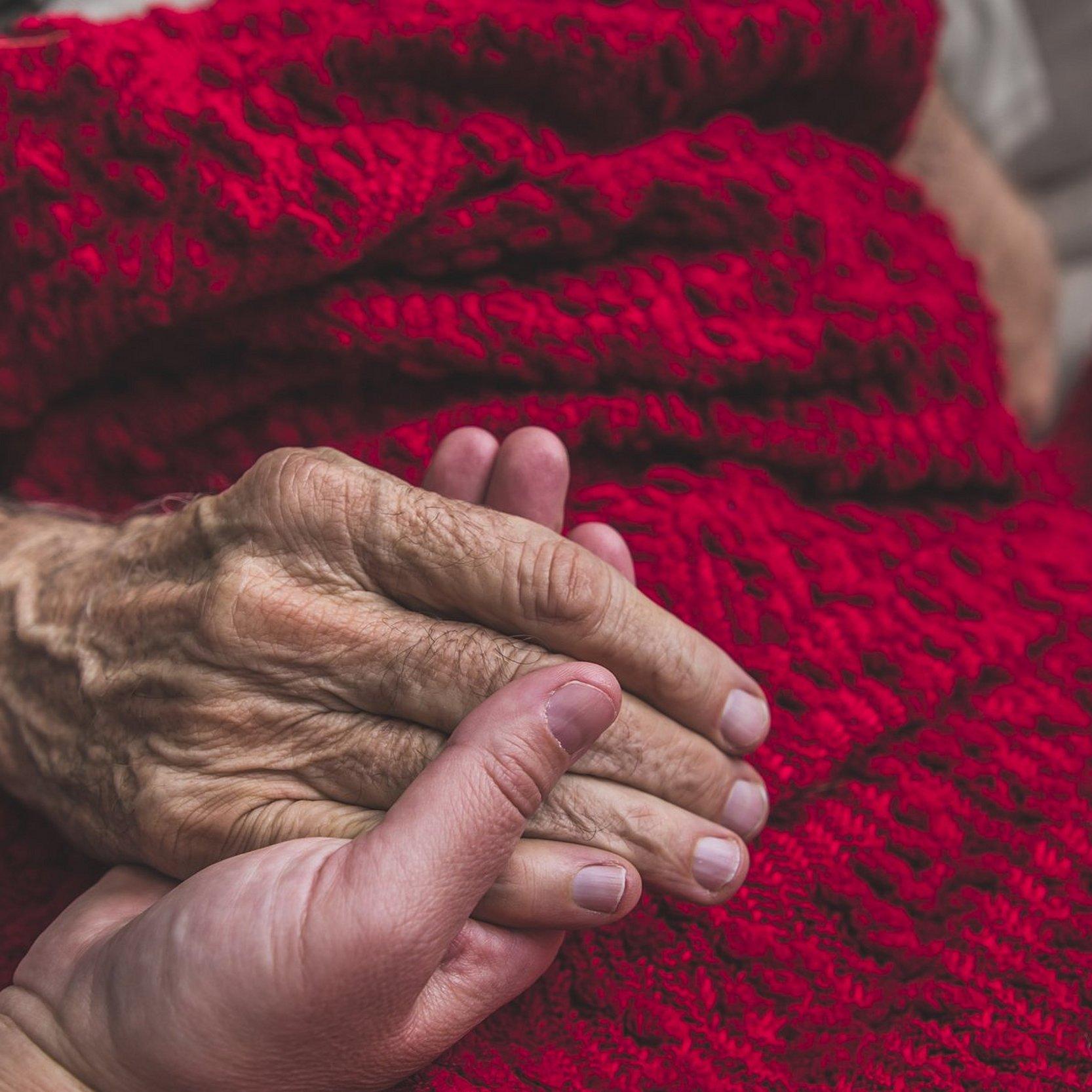 Eine junge Hand hält die alte Hand einer Sterbenden. Diese ist mit einer schönen, roten Decke zugedeckt.