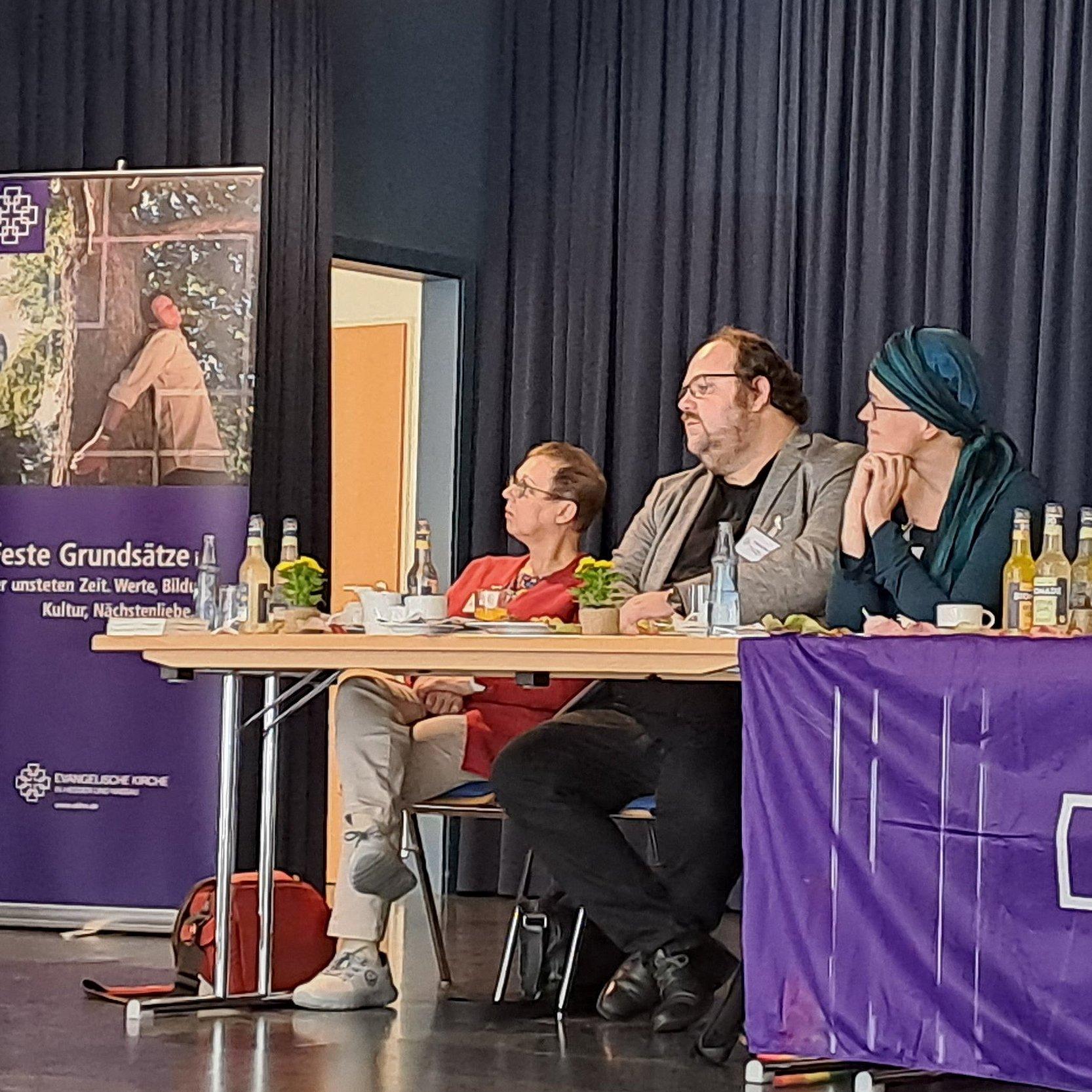 Auf einem Podium steht links eine Frau am Rednerpult. Rechts sitzen mehrere Personen hinter einem Tisch Das Podium ist geschmückt mit lila Banner der Evangelischen Kirche in Hessen und Nassau.