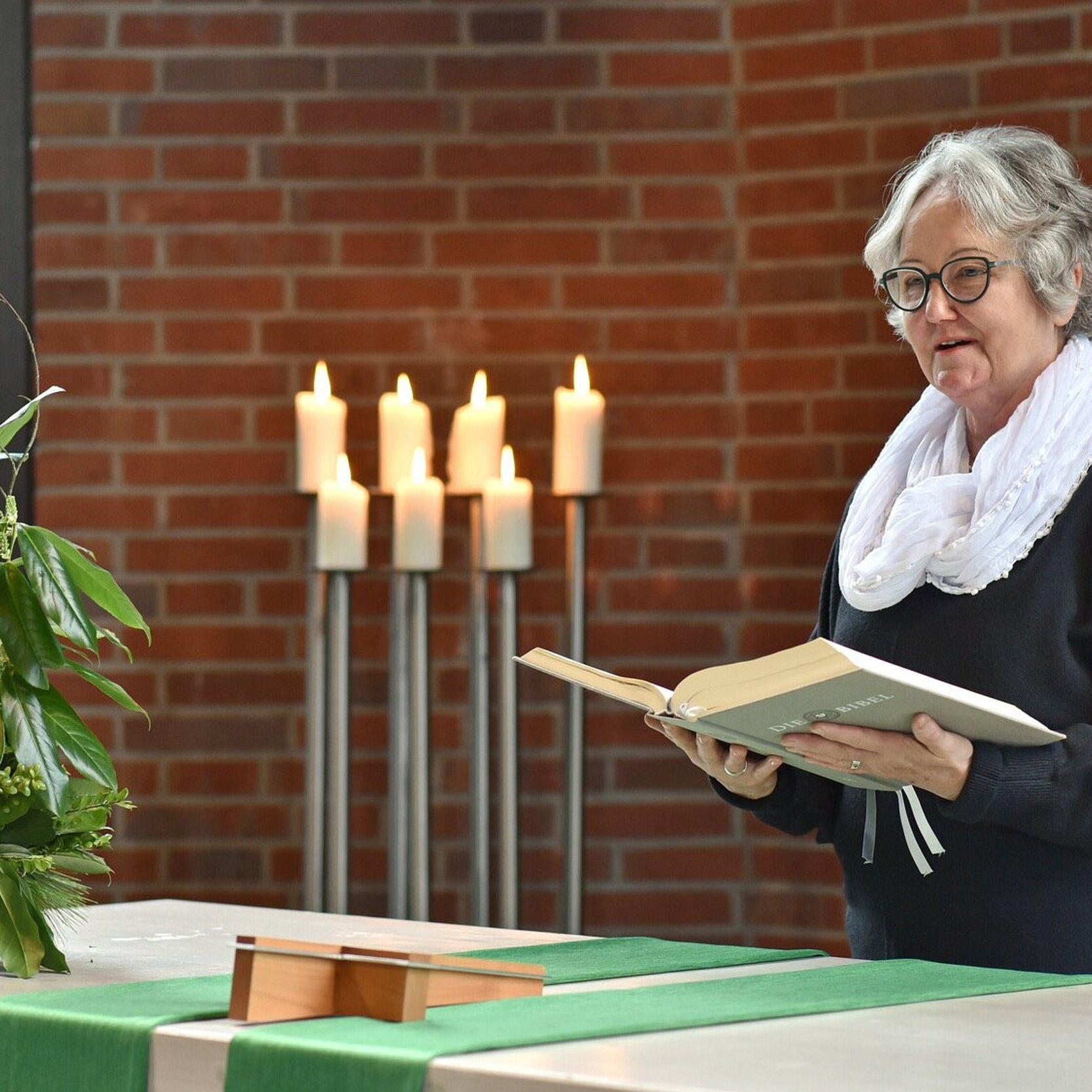 Ein predigende Frau steht hinter einem Altar mit Blumen und liest aus einer großen Bibel, die sie in Händen hält. Im Hintergrund brennen sieben Kerzen auf hohen silbernen Lauchtern.