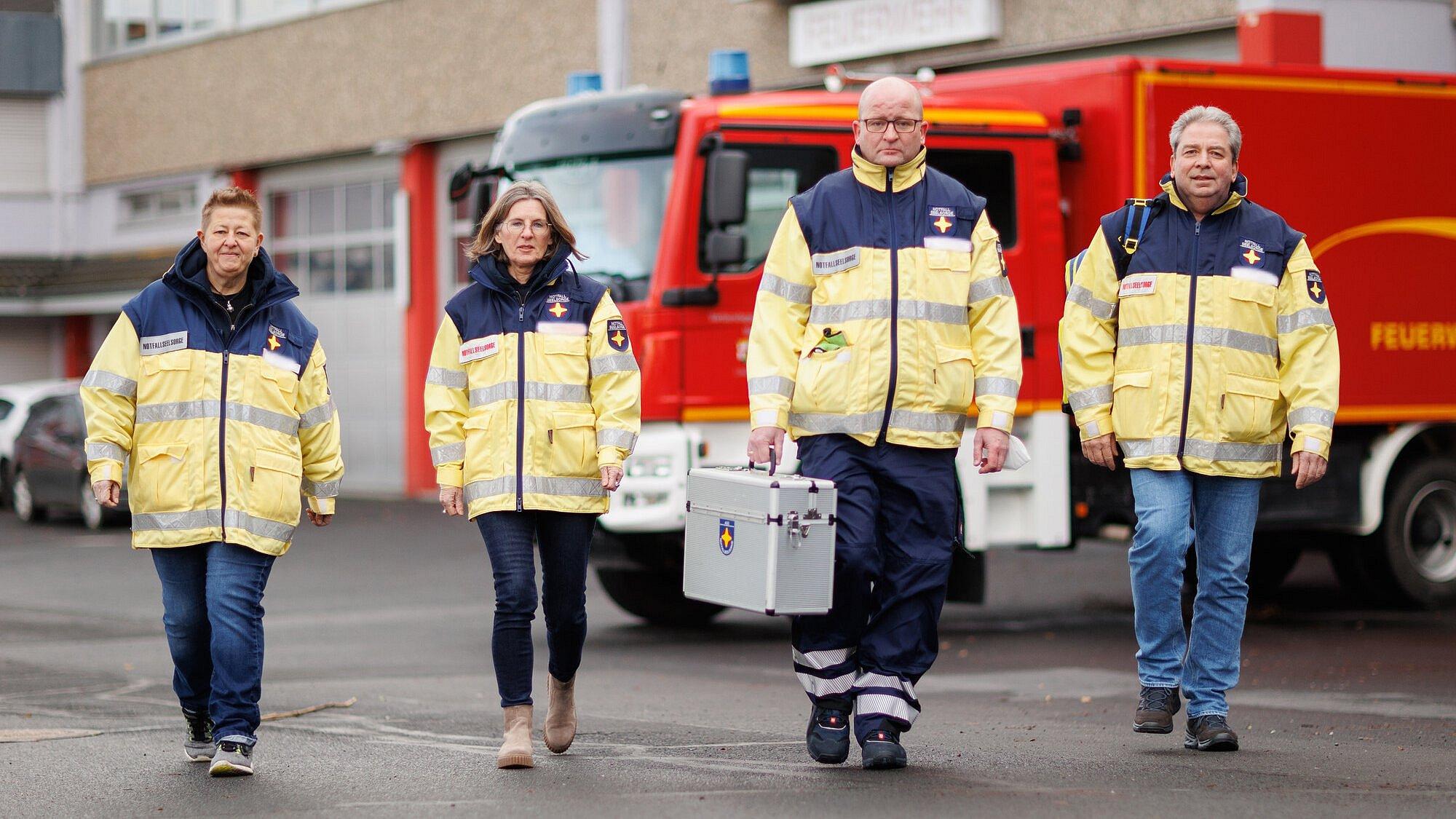 Vier Personen laufen auf den Fotografen zu. Sie tragen Uniformen der Notfallseelsorge. Im Hintergrund steht ein Feuerwehrfahrzeug vor der Brandwache.
