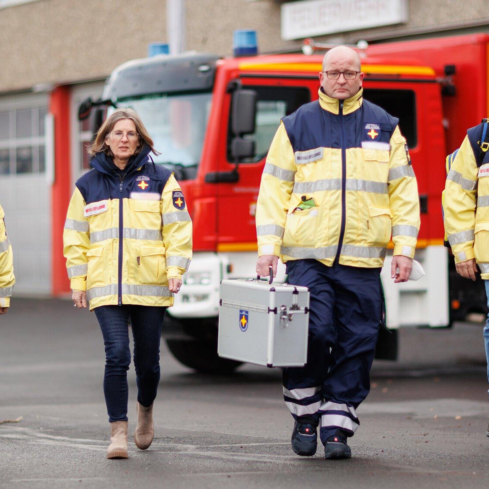Notfallseelsorge im Einsatz Vier Personen laufen auf den Fotografen zu. Sie tragen Uniformen der Notfallseelsorge. Im Hintergrund steht ein Feuerwehrfahrzeug vor der Brandwache.