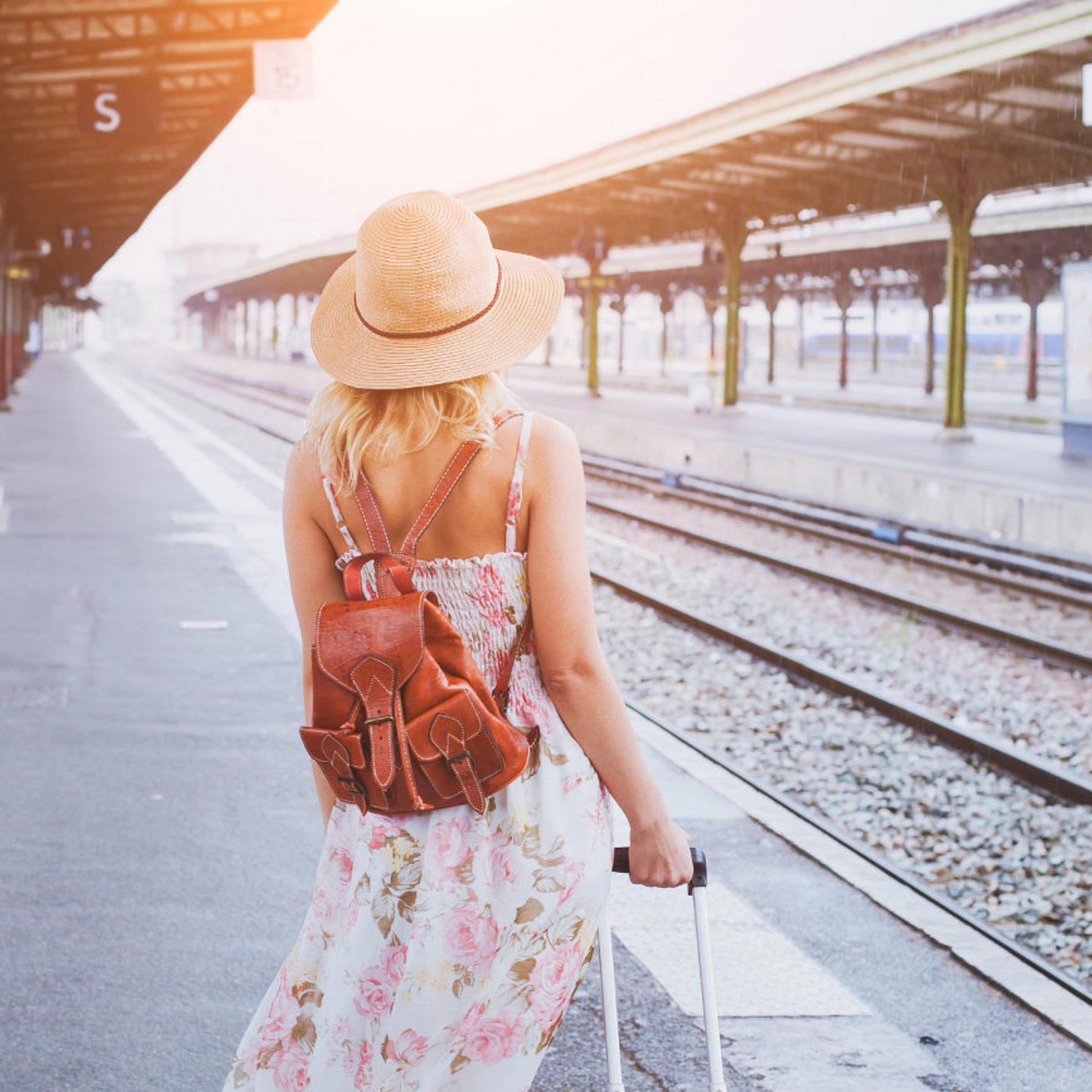 Frau steht mit einem Sommerkleid und Koffer und Rucksack auf dem Bahnhof am Gleis  und schaut in die Ferne.