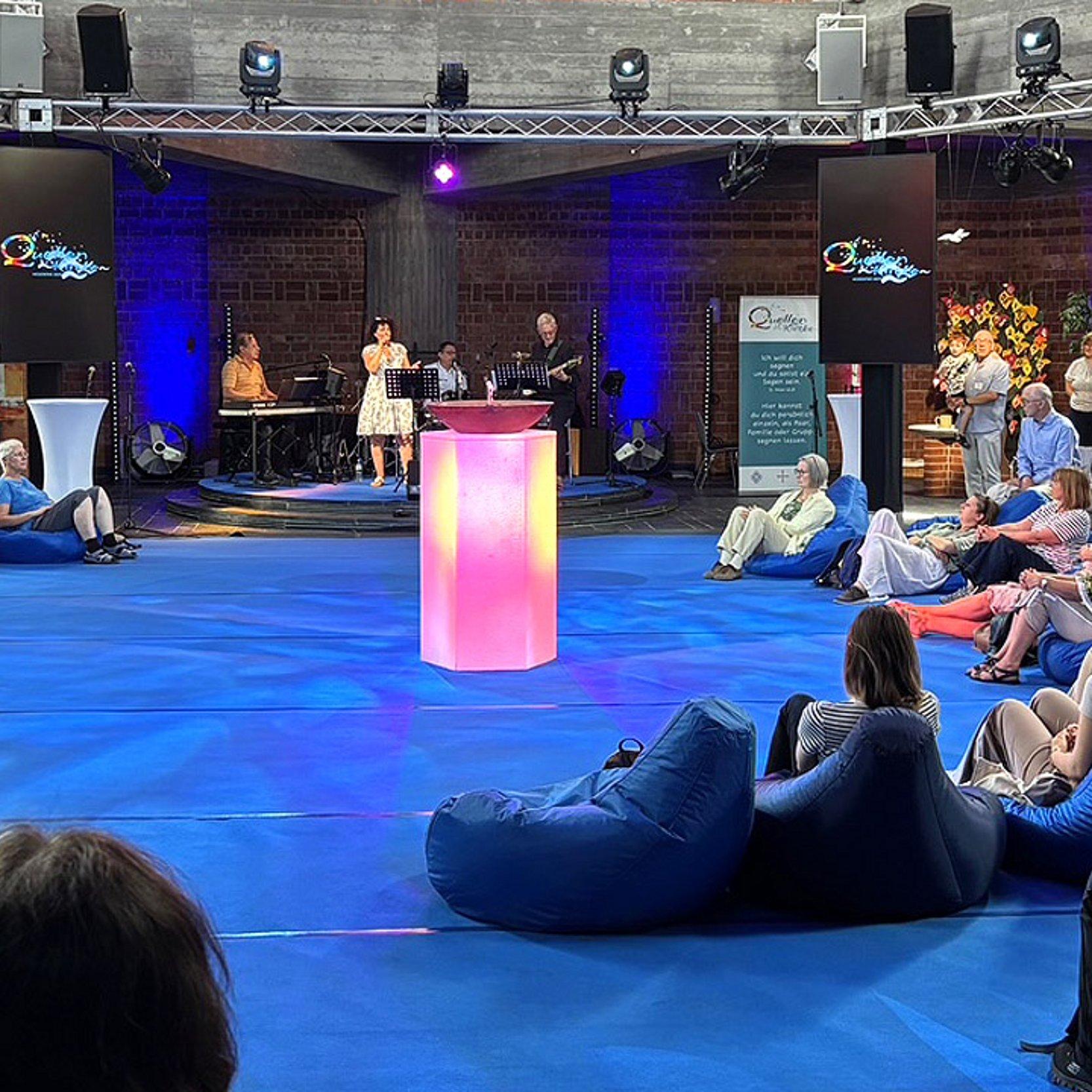 Quellenkirche Menschen auf Bänken und Loungesesseln rund um den rötlich beleuchteten Pult mit der kleinen Wasserfontaine