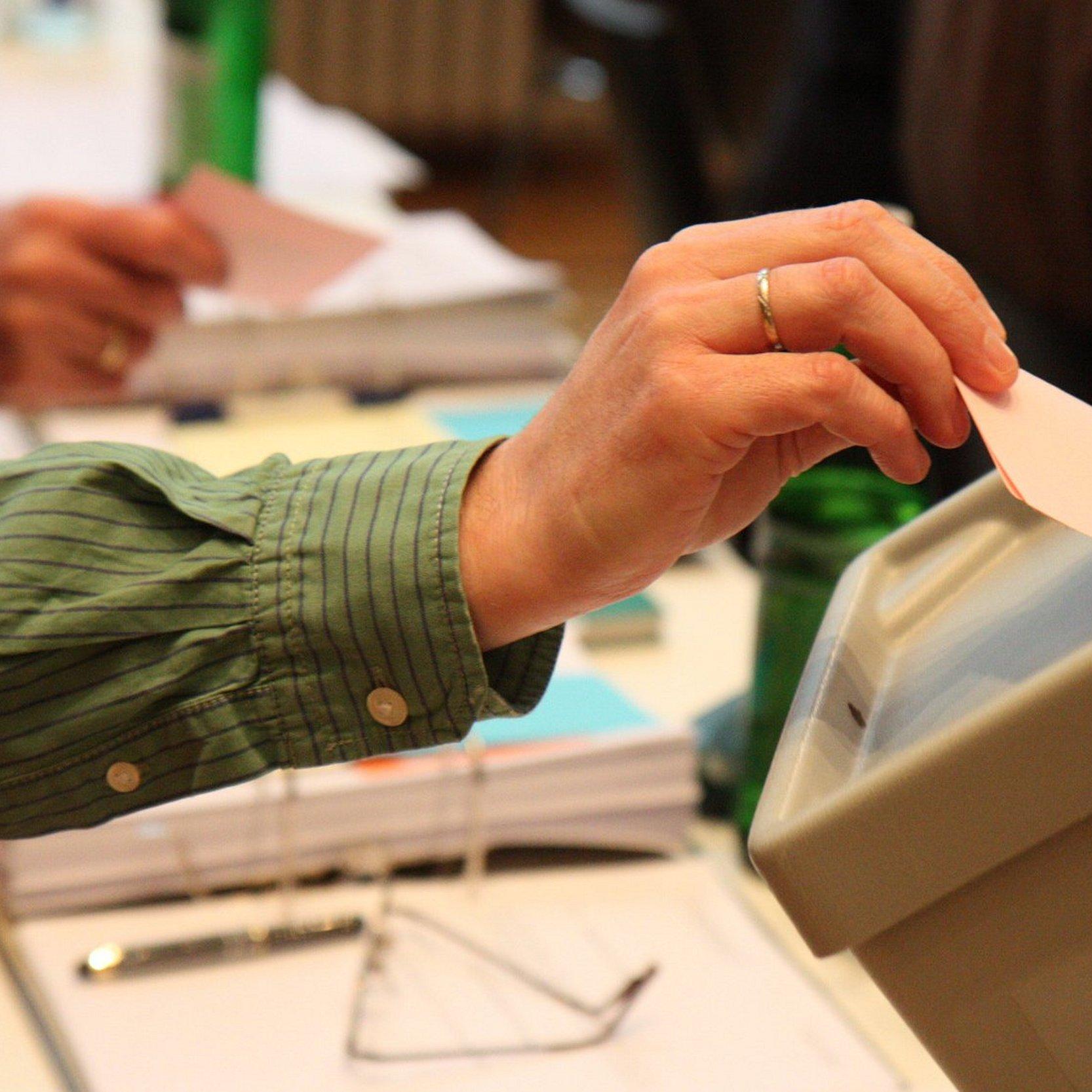 Eine Hand steckt einen Wahlzettel in eine Wahlurne mit einem Logo der EKHN..