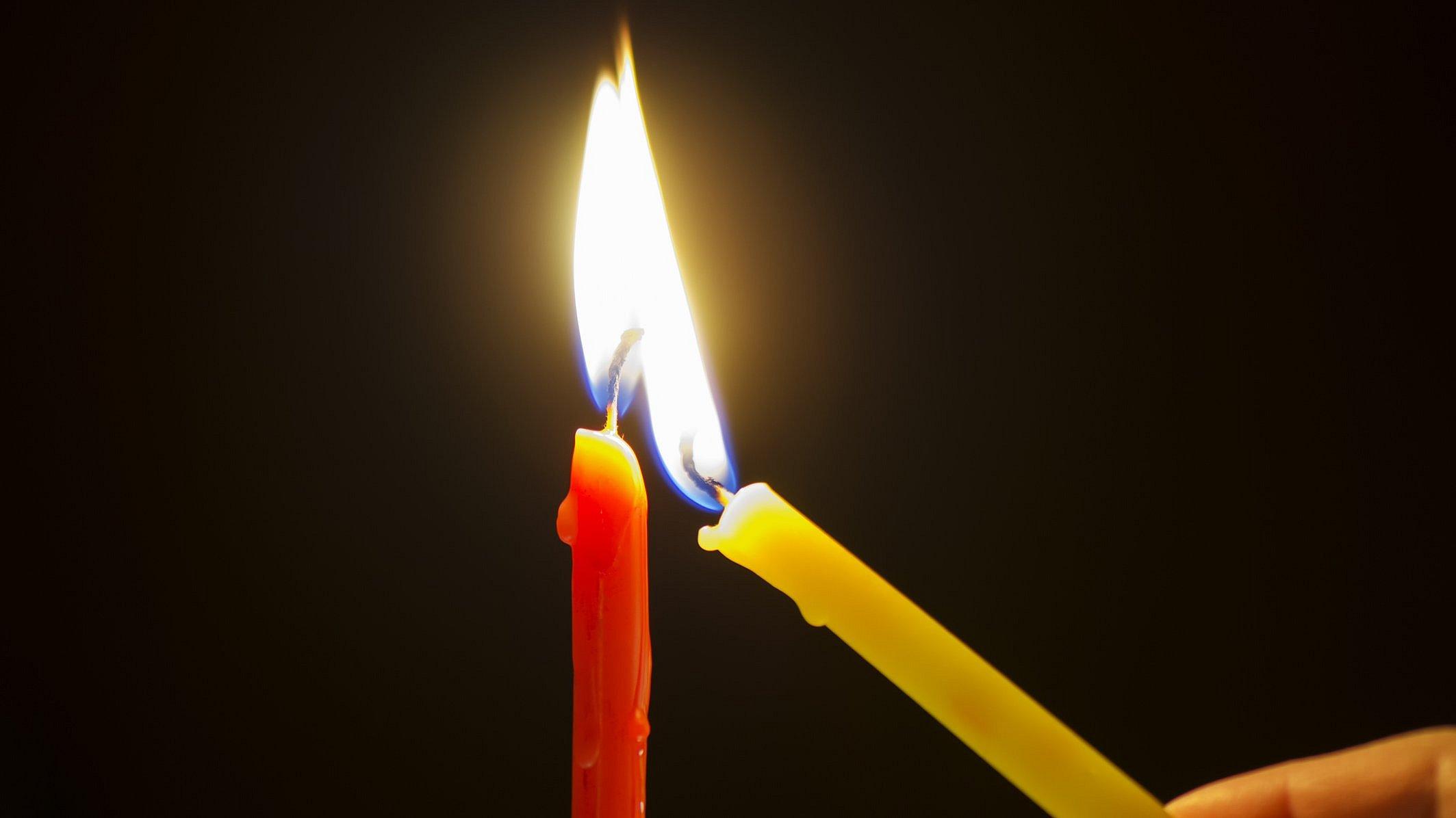 Eine gelbe schmale Kerze wird an einer roten schmalen Kerze angezündet.