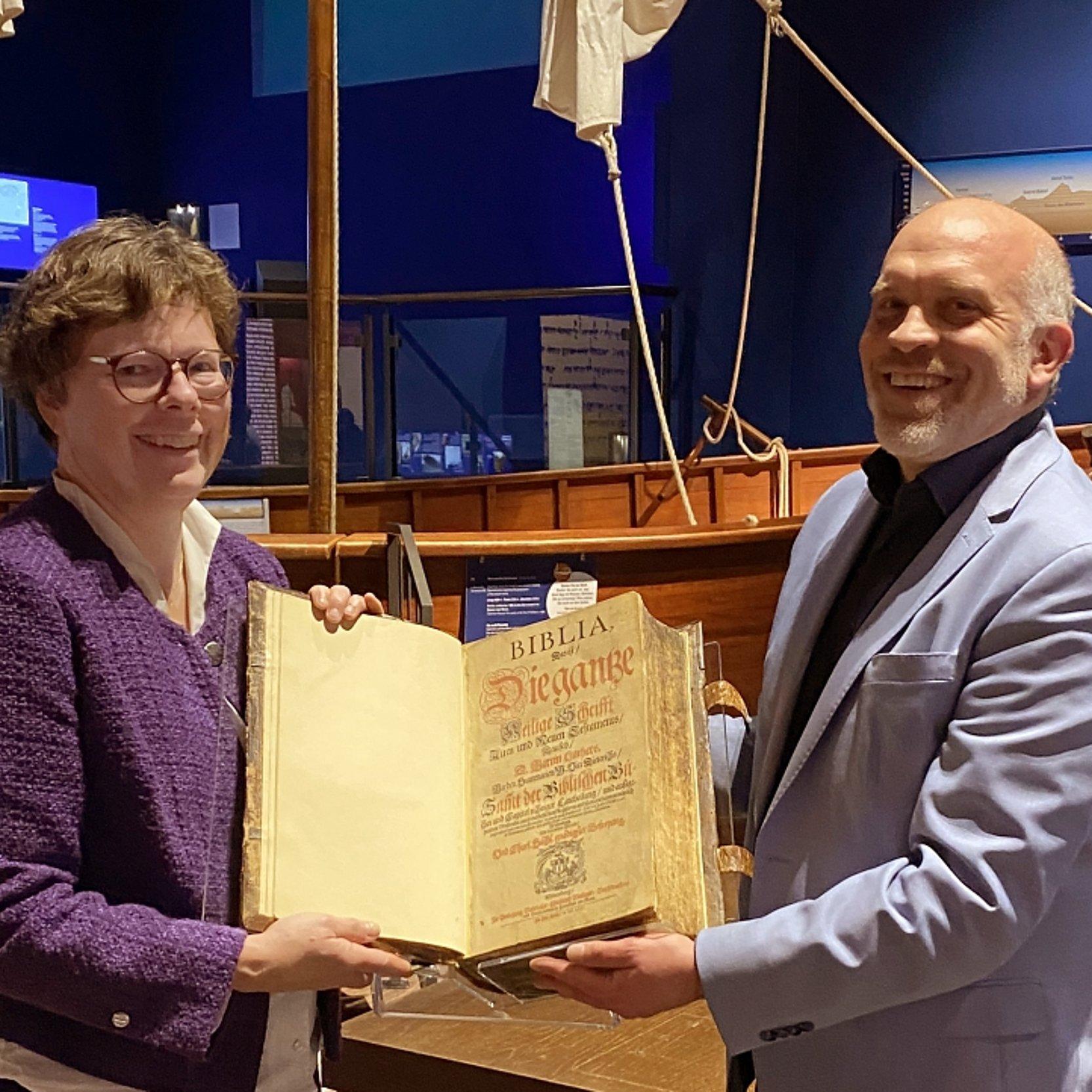 Bischöfin Hofman Veit Dinkelaker Wust-Bibel Bischöfin Hofmann übergibt die historische Bibel an Museumsdirektor Veit Dinkelaker. Im Hintergrund ist ein historisches Segelschiff im Austellungsraum des Museums zu sehen.