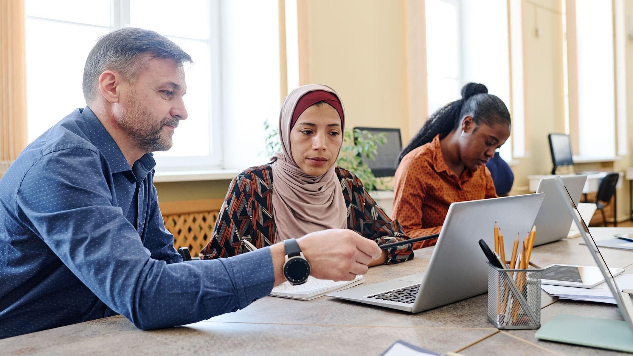 Ein Mann erklärt einer Frau mit Migrationshintergrund eine Aufgabe an einem Computer