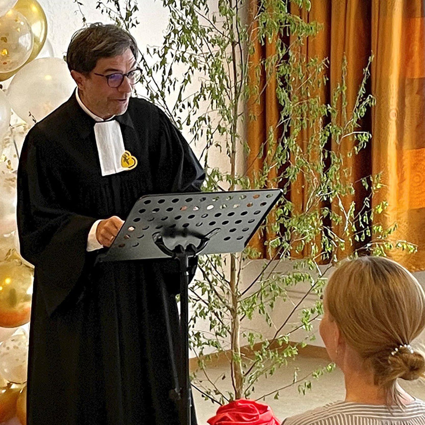 Ein Pfarrer steht vor einem jungen Paar. Dahinter ist eine Girlande mit goldenen und weißen Luftballons.