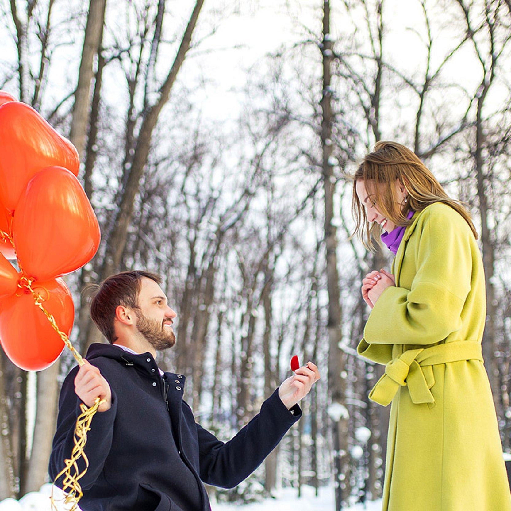 Verlobung Ein Mann geht vor einer Frau in die Knie, er hat viele rote Herz-Luftballons dabei, es ist draußen im Schnee.