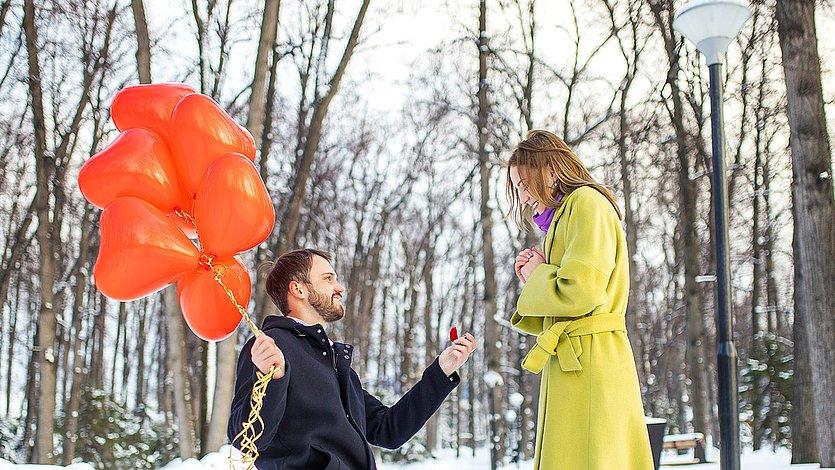 Verlobung Ein Mann geht vor einer Frau in die Knie, er hat viele rote Herz-Luftballons dabei, es ist draußen im Schnee.