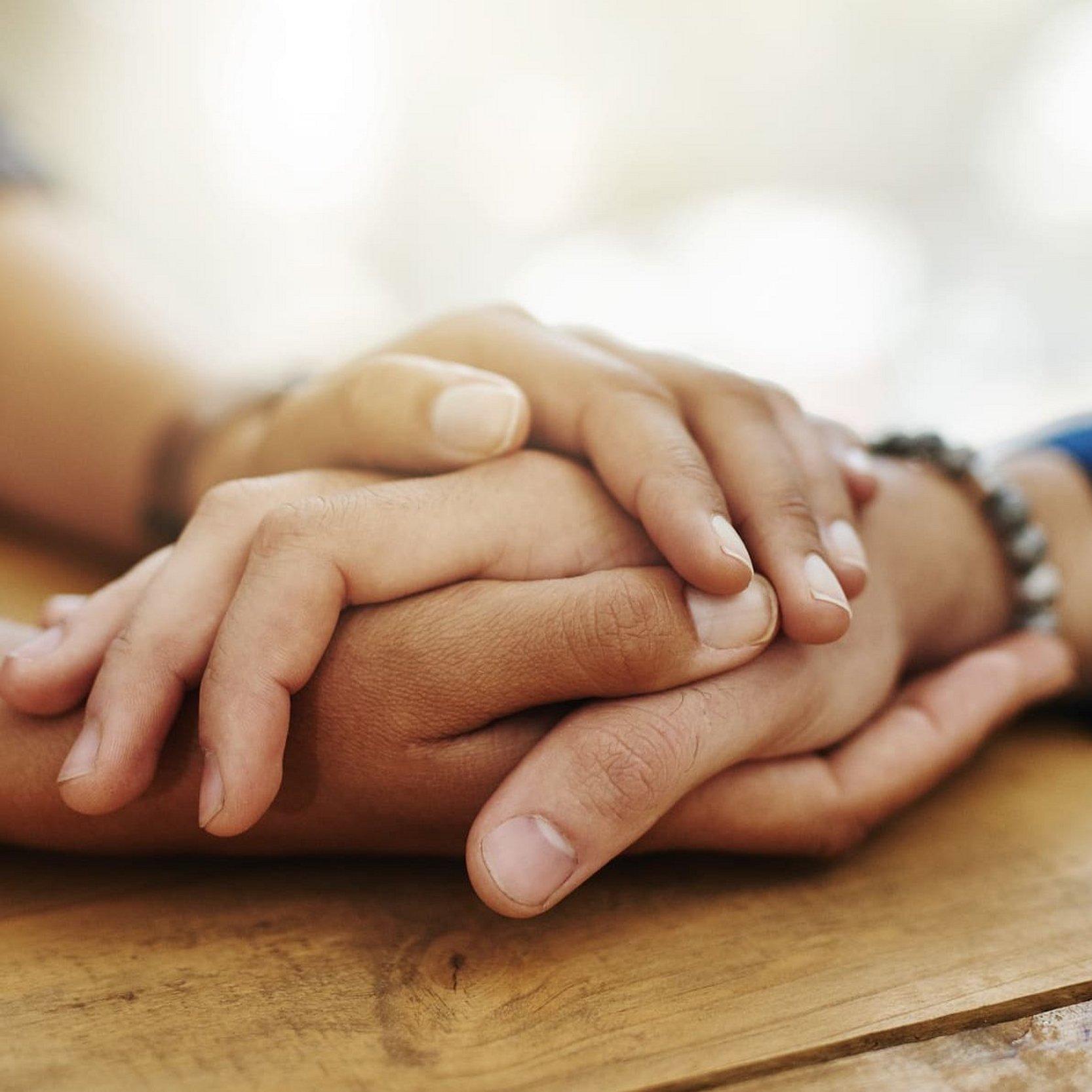 Diakonie Hessen Helfen aus Menschlichkeit Vier auf dem Tisch ruhende Hände von zwei Menschen halten sich gegenseitig.