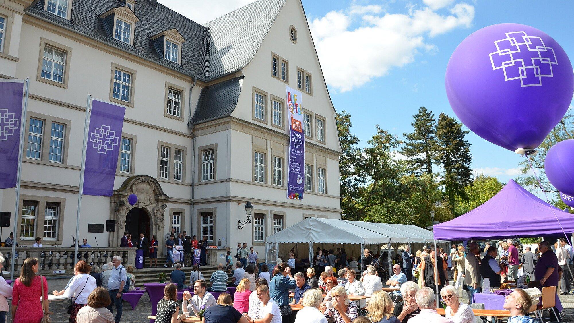 FUNDUS_7763_01_Paulusplatz Vor dem historischen Gebäude der Kirchenverwaltung der EKHN in Darmstadt findet eine öffentliche Veranstaltung im Freien statt. Zahlreiche Menschen sitzen an Biertischgarnituren oder stehen auf dem Platz. Lila Fahnen, Luftballons und ein Pavillon mit dem EKHN-Logo prägen die Szenerie.