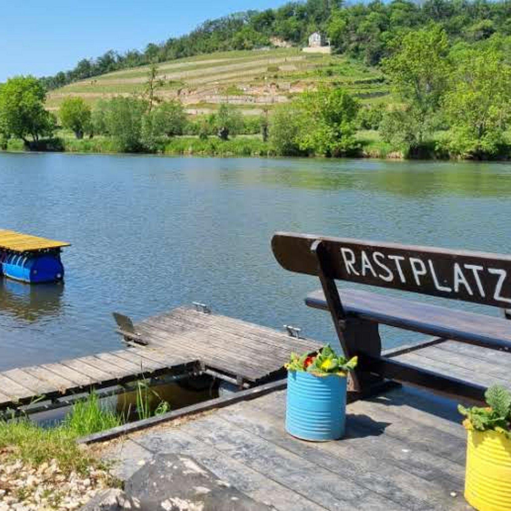 Rastplatz Eine Ruhebank am Flussufer mit der Aufschrift "Rastplatz". vor der Ruhebank stehen ein gelber und ein blauer Pflanzkübel mit Blumen. Auf der gegenüberliegenden Seite des Flusses sind Bäume und grüne Sträucher zu erkennen. Im Hintergrund ein wolkenloser, blauer Himmel.