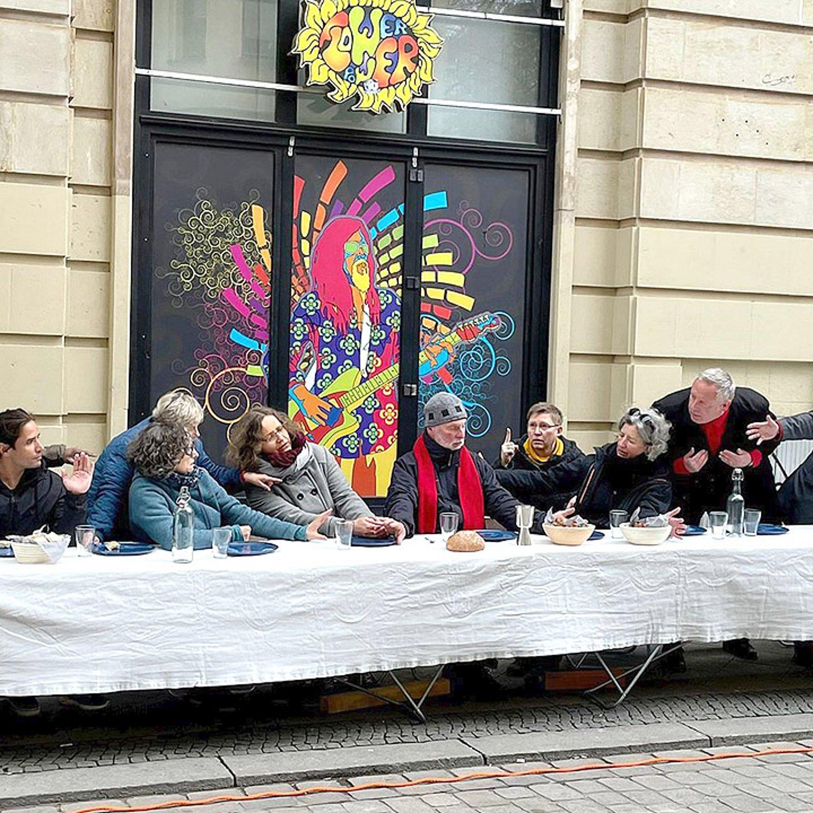 Menschen in Alltagskleidung an einem langen Tisch auf einem Bürgersteig