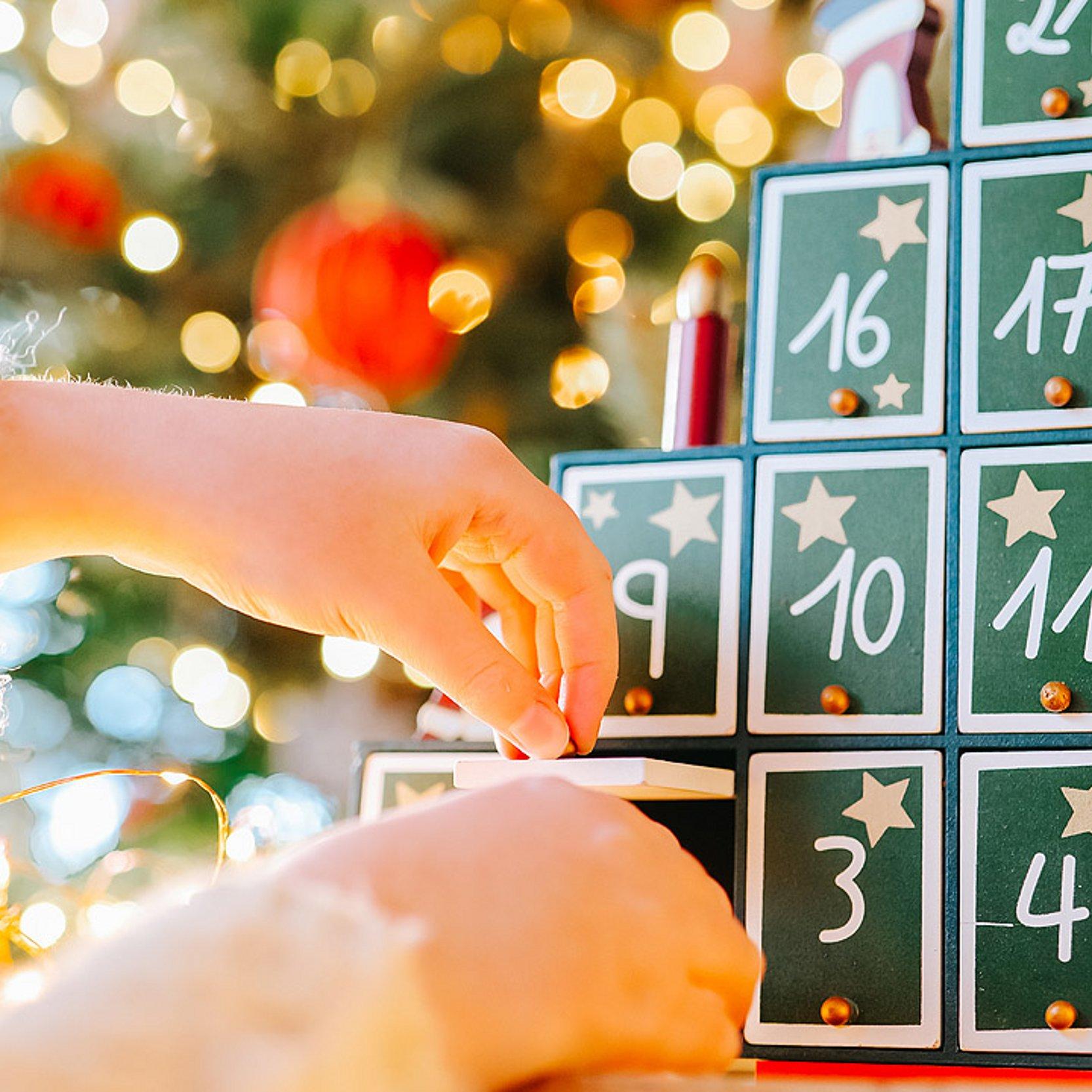 Adventskalender Die Hand eines Mädchen öffnet eine kleine Schublade eines bemalten Adventskalenders aus Holz
