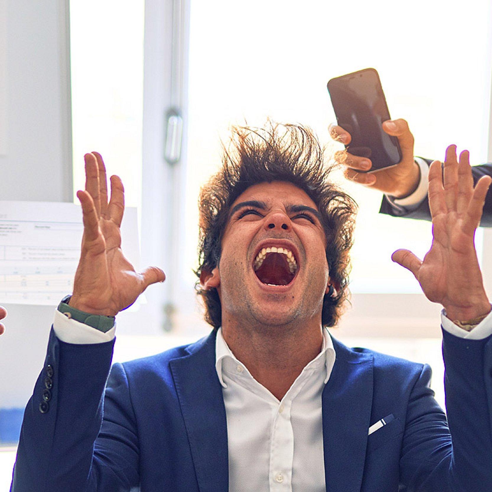 Stress und Zeitdruck im Job Einem Mann stehen die Haare zu Berge, ihm werden Blätter mit Diagrammen und Wecker entgegengestreckt