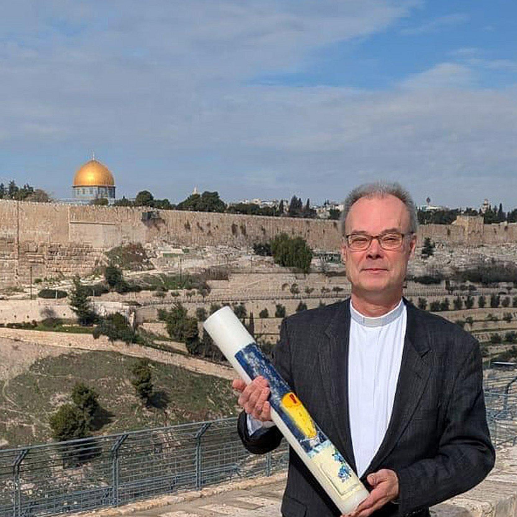 Andreas Goetze in Jerusalem Ein Mann im dunklen Anzug trägt eine große Kerze vor der Stadtansicht Jerusalems mit Felsendom
