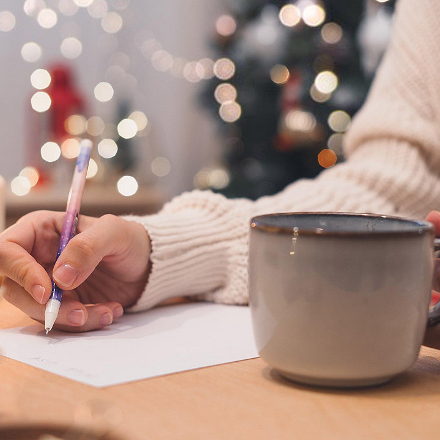Rauhnächte Hand einer Frau die schreibt und eine große Tasse mit einem heißen Getränk. Dahinter Lichter und Kerzen, die noch auf Weihnachten hindeuten