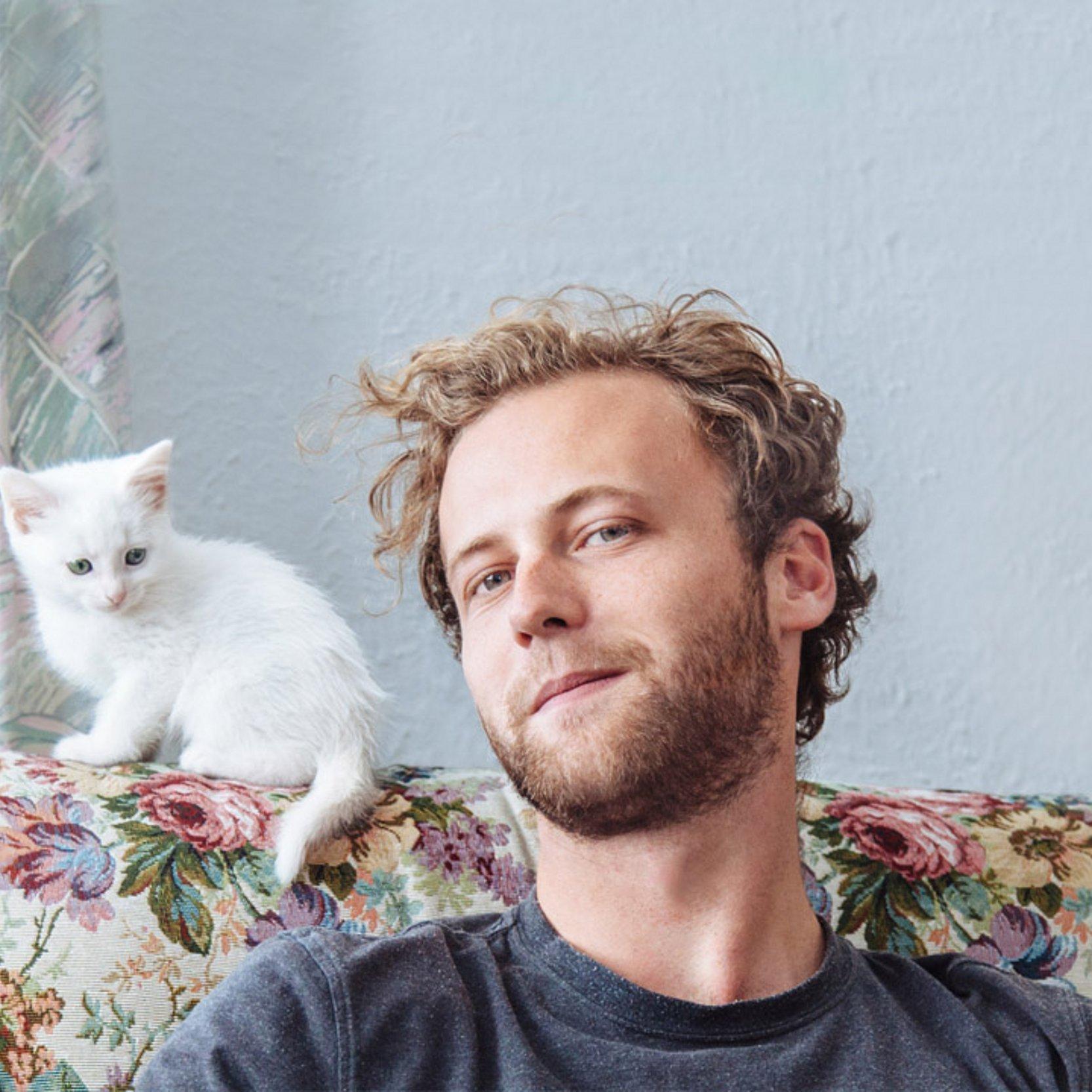 ohne Härte Junger Mann sitzt auf einem Sessel mit Blumenmuster, auf der Lehne sitzt eine junge, weiße Katze