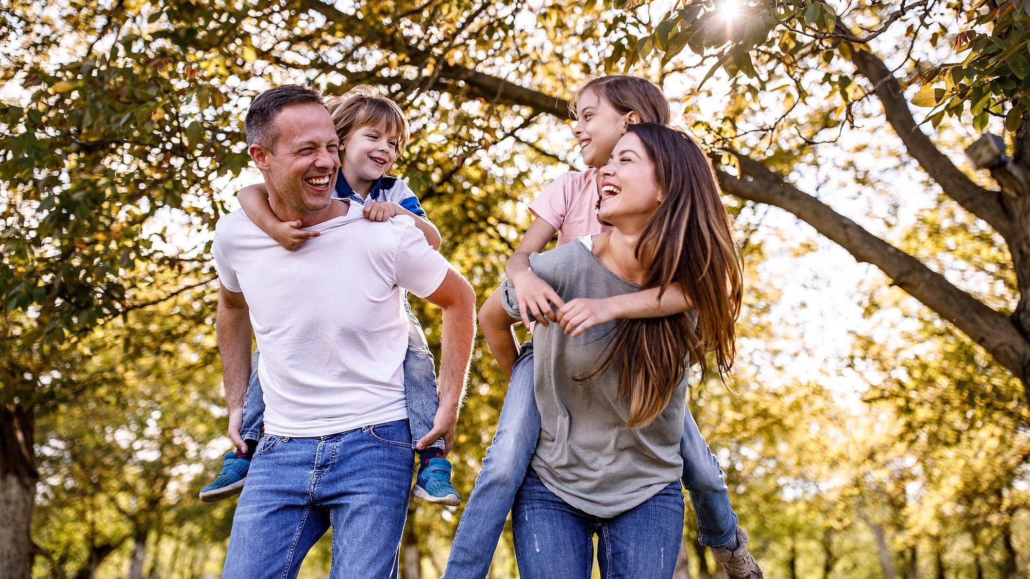 Familie hat Spaß zusammen und wandert Ein Vater und eine Mutter gehen durch den Wald, sie lachen und haben jeweils ein Kind auf ihrem Rücken. Die Kinder lachen und schauen sich an. Alle sind glücklich beim Huckepack-Spiel.