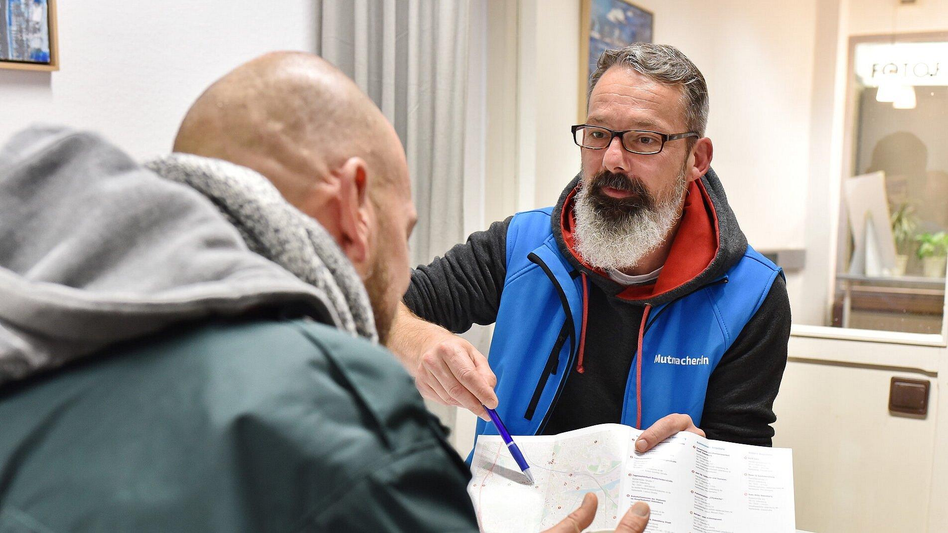 Mitarbeiter der Bahnhofsmission im Beratungsgespräch. Mitarbeiter zeigt Klienten eine Broschüre oder Karte.