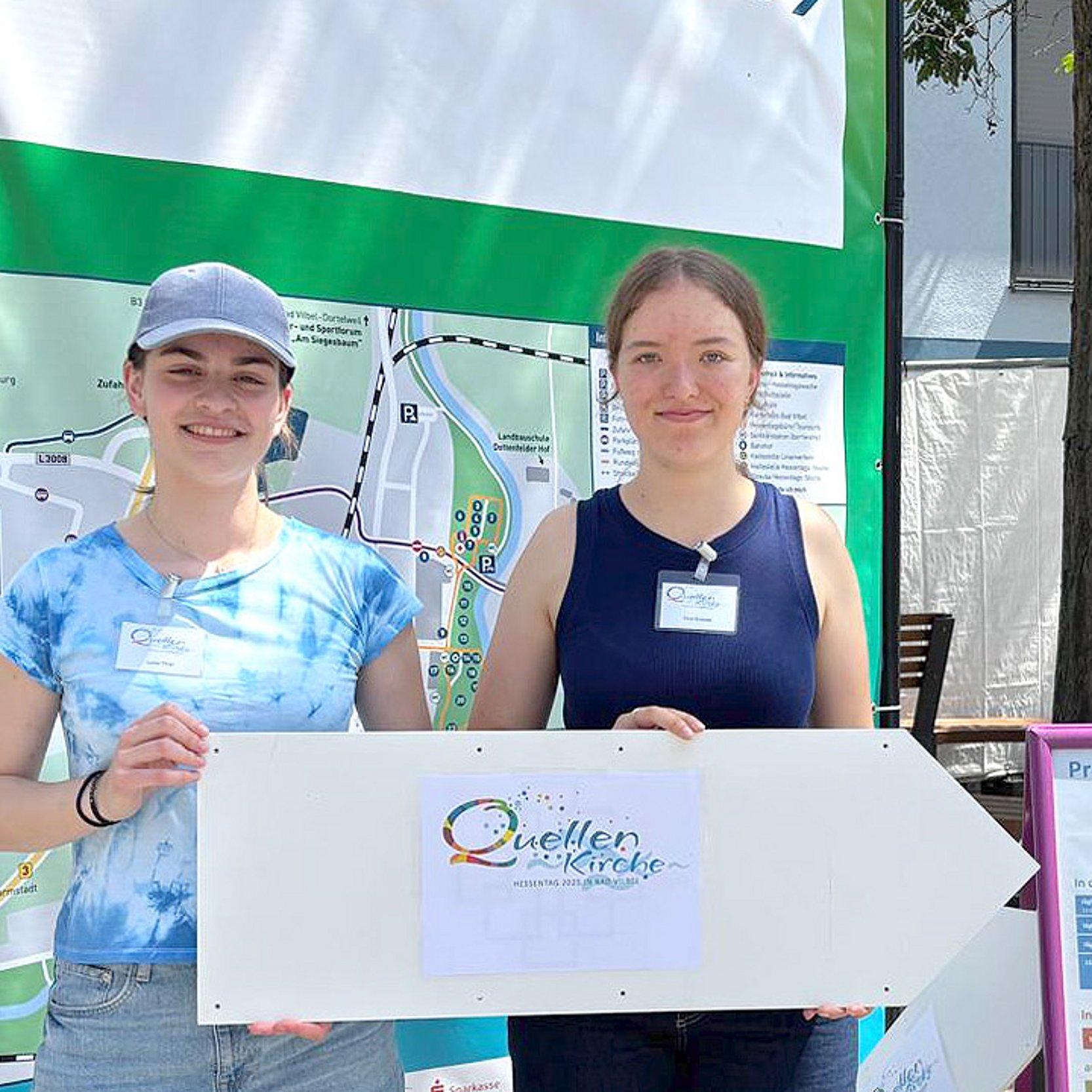 Quellenkirche Zwei junge Frauen halten ein Schild mit der Aufschrift "Quellenkirche"