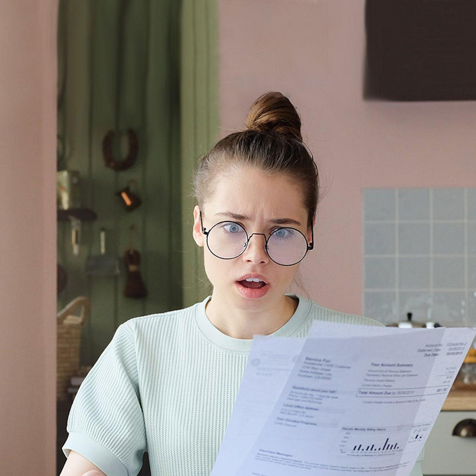 Schulden Frau mit High Bun schaut entsetzt auf ein Papier und ihr Smartphone