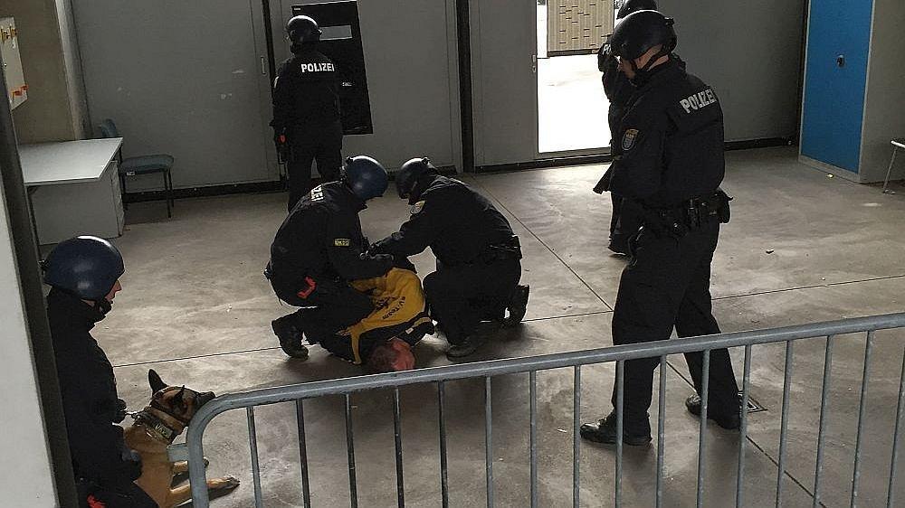 Polizisten in Schutzkleidung nehmen eine am Boden liegende Person fest.