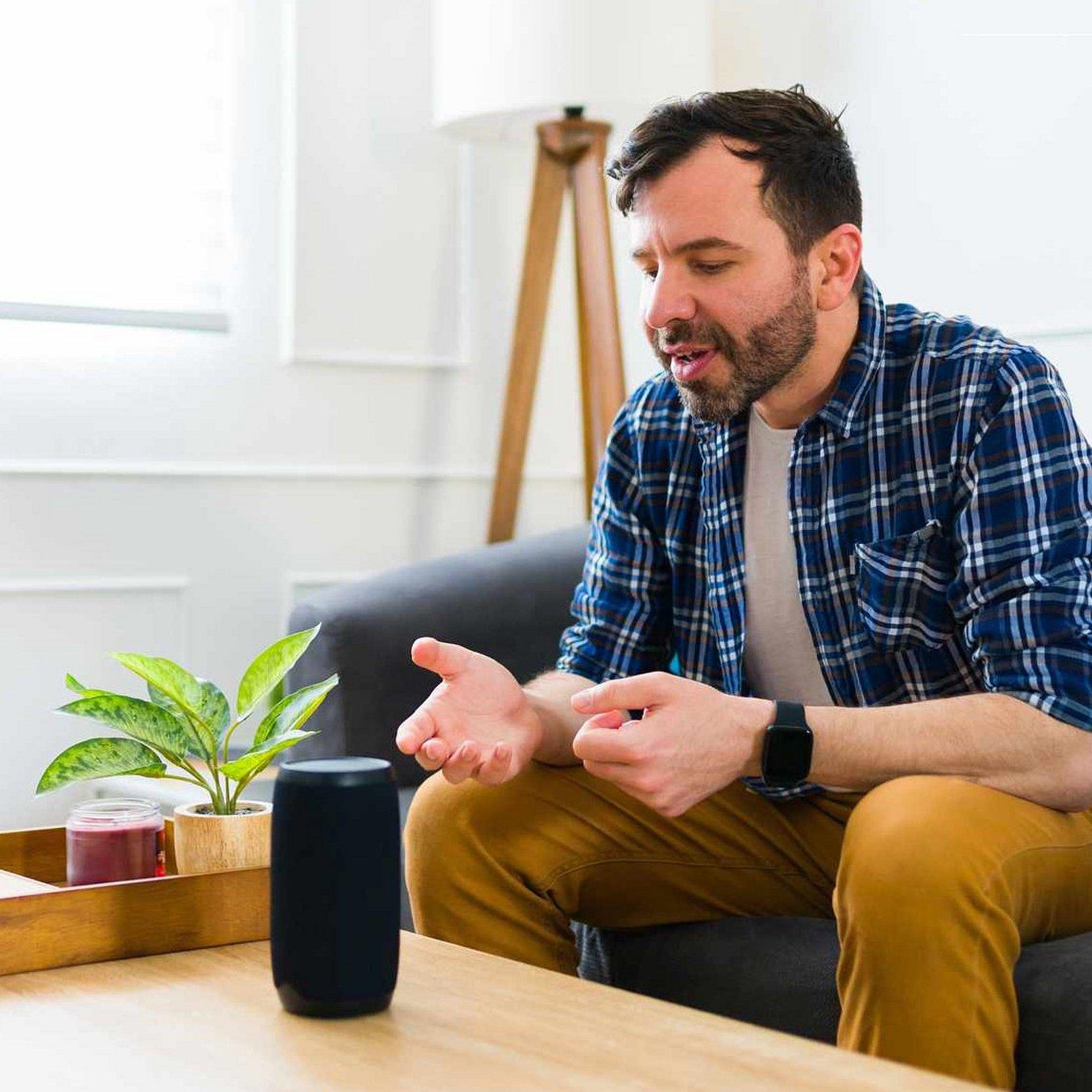 Ein Mann sitzt auf dem Sofa und Sprachassistentin Alexa steht auf dem Tisch vor ihm. Der Mann spricht zu Alexa.