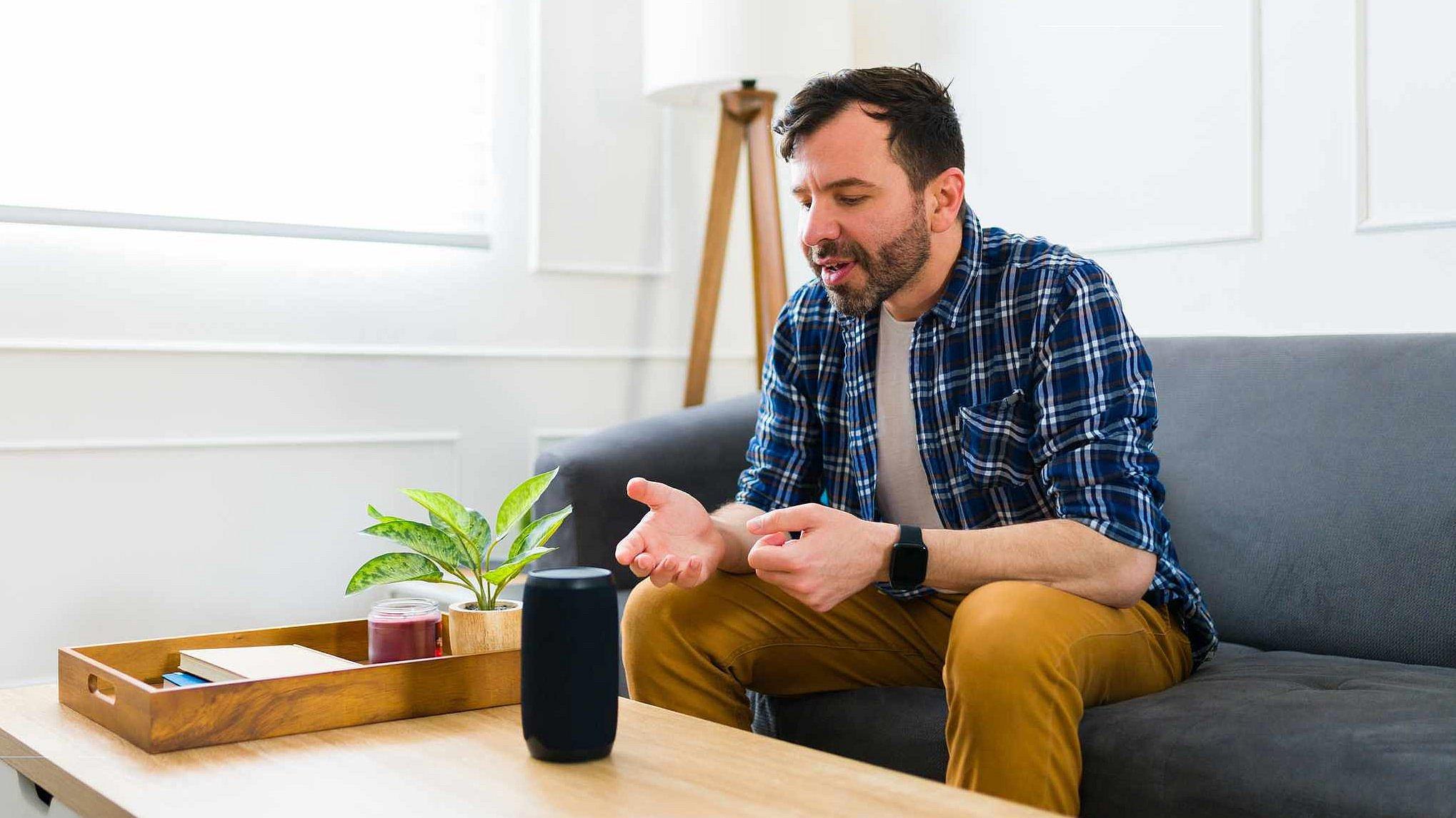 Ein Mann sitzt auf dem Sofa und Sprachassistentin Alexa steht auf dem Tisch vor ihm. Der Mann spricht zu Alexa.
