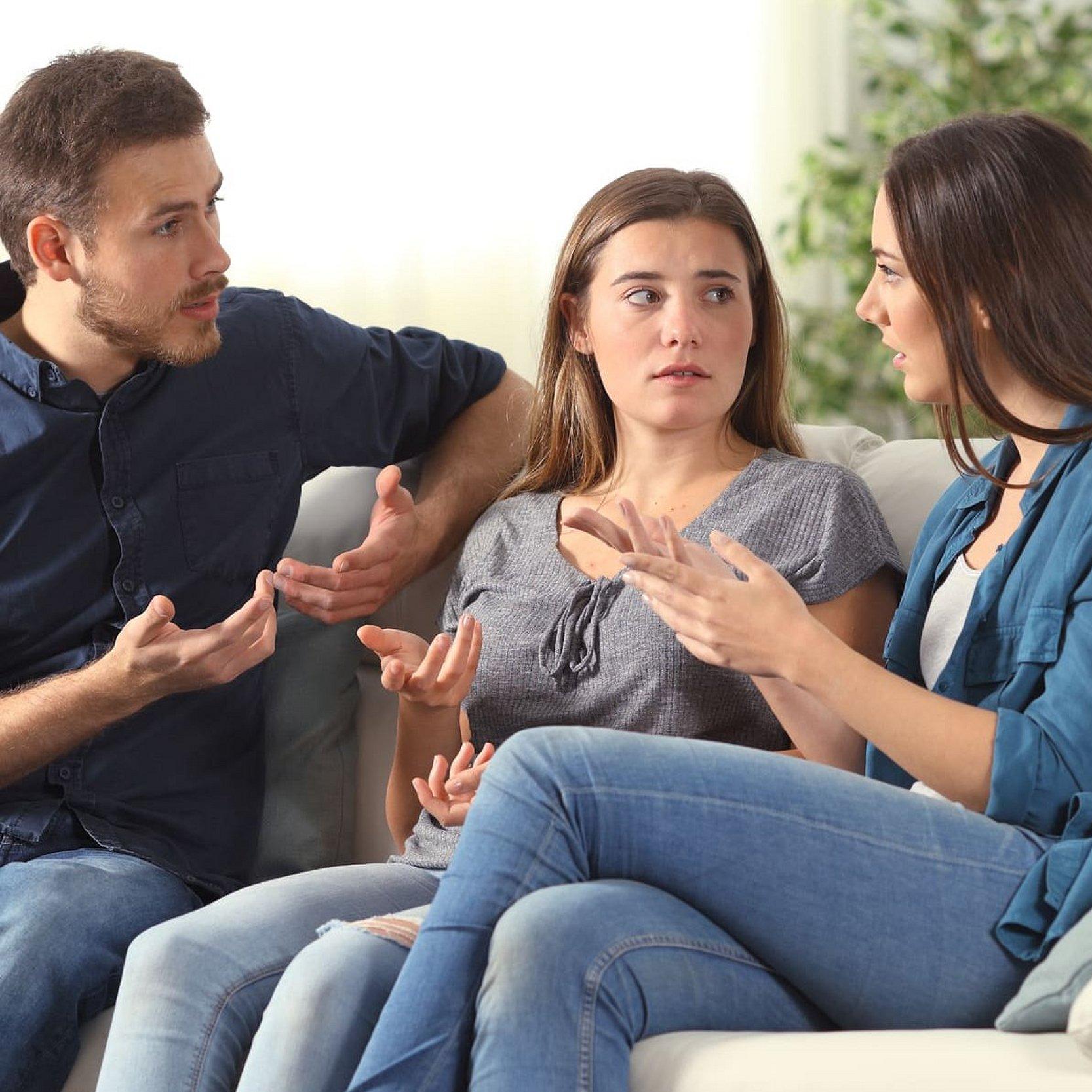 Verschwörungsideelogien_Diskussion_Demokratie Ein junger Mann und zwei junge Frauen sitzen auf einer weißen Couch und diskutieren lebhaft miteinander, sie gestikulieren dabei.