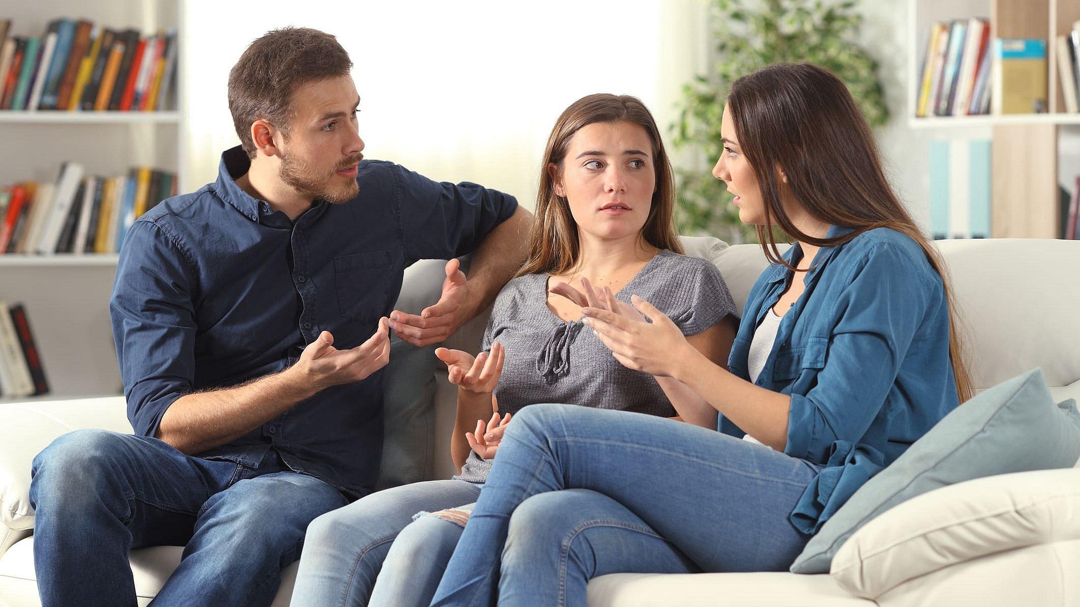 Ein junger Mann und zwei junge Frauen sitzen auf einer weißen Couch und diskutieren lebhaft miteinander, sie gestikulieren dabei.