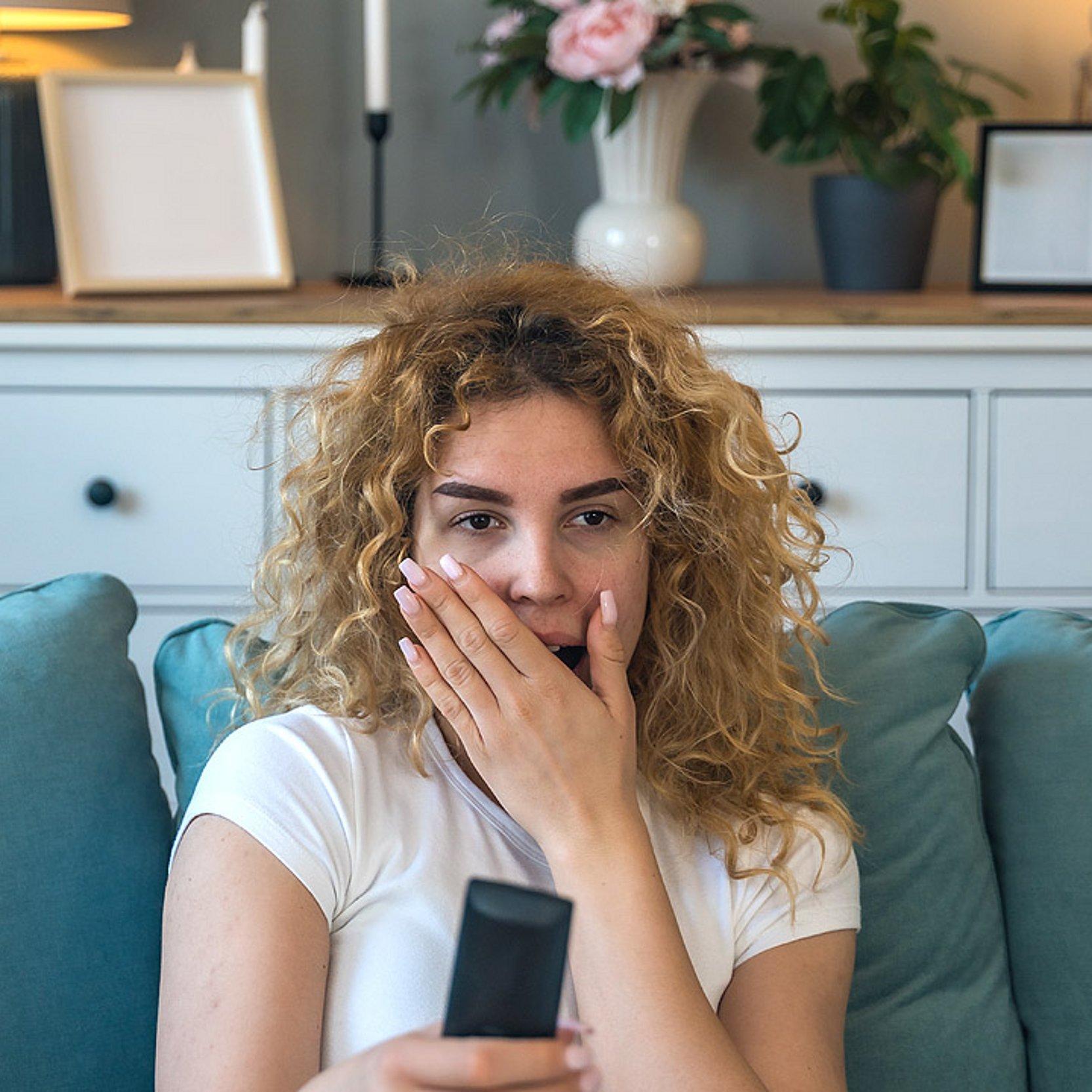 Erschrocken über die aktuelle Nachrichtenlage Eine Frau auf der Couch mit Fernbedienung ist erschrocken über die aktuelle Nachrichtenlage