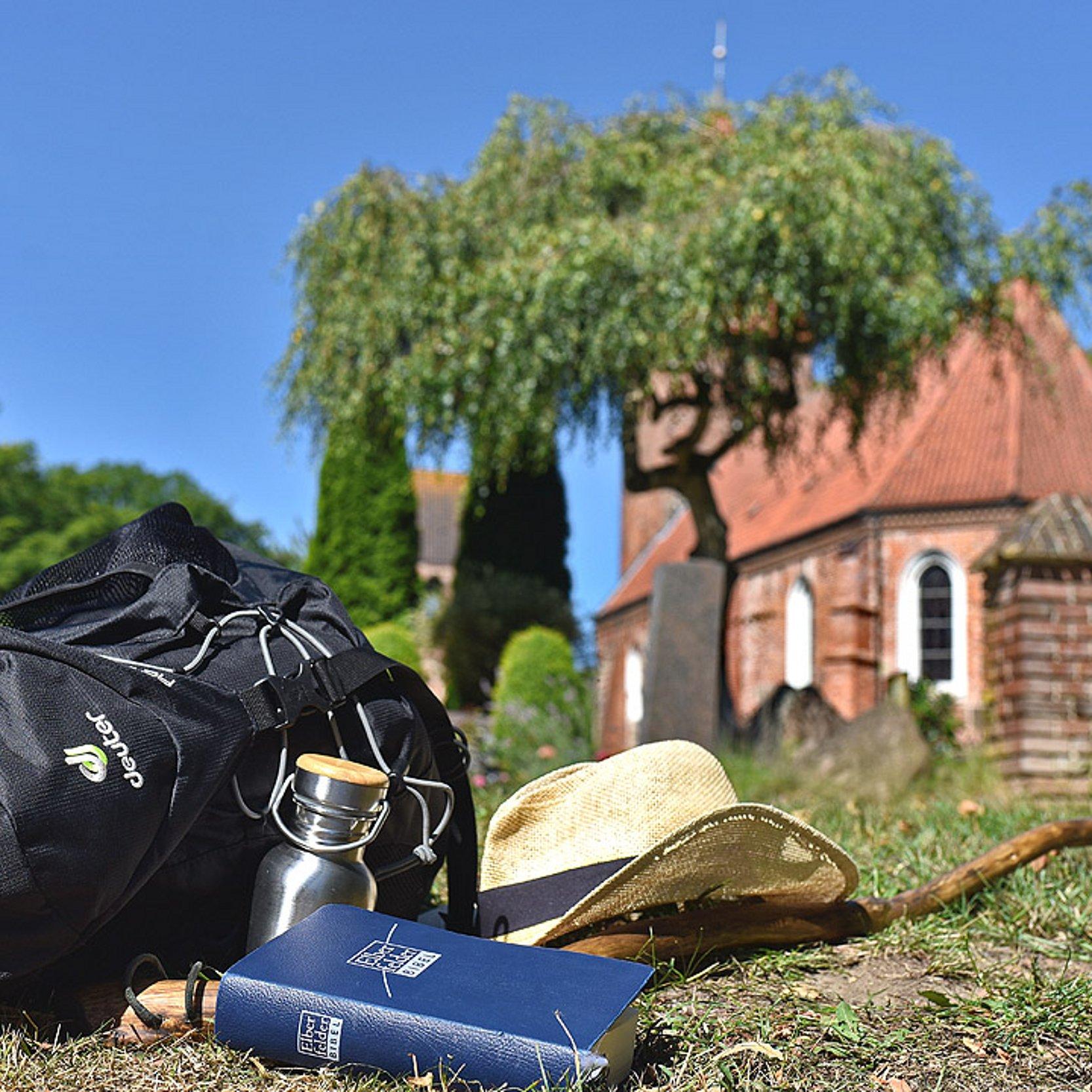 Pilgerrucksack Ein Rucksack, ein Hut und eine Trinkflasche liegen auf dem Gras vor einer kleinen Kirche