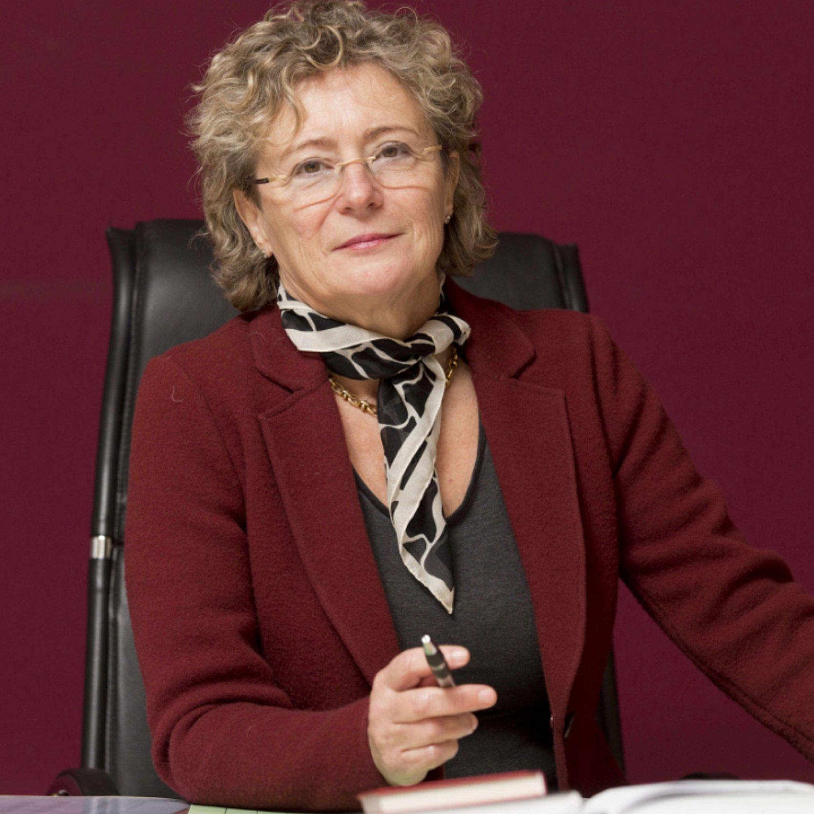 Esther Gebhardt Esther Gebhardt mit rotem Blazer und buntem Halstuch sitzt an ihrem Schreibtisch und hält einen Kugelschreiber in der Hand