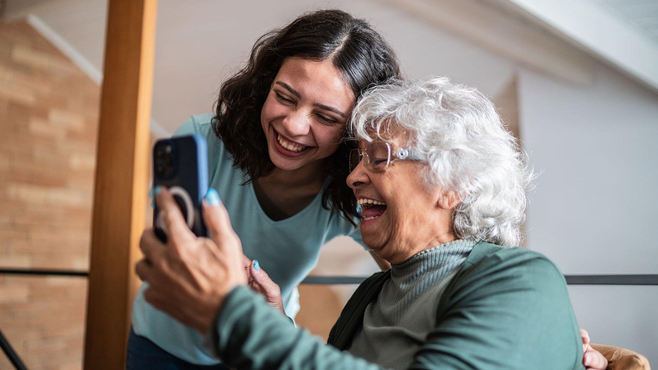 Eine ältere Frau sitzt und hält ein Smartphone in der Hand, eine junge Frau beugt sich über sie und schaut auch in das Smartphone, beide lächeln und sehen sehr freundlich aus.