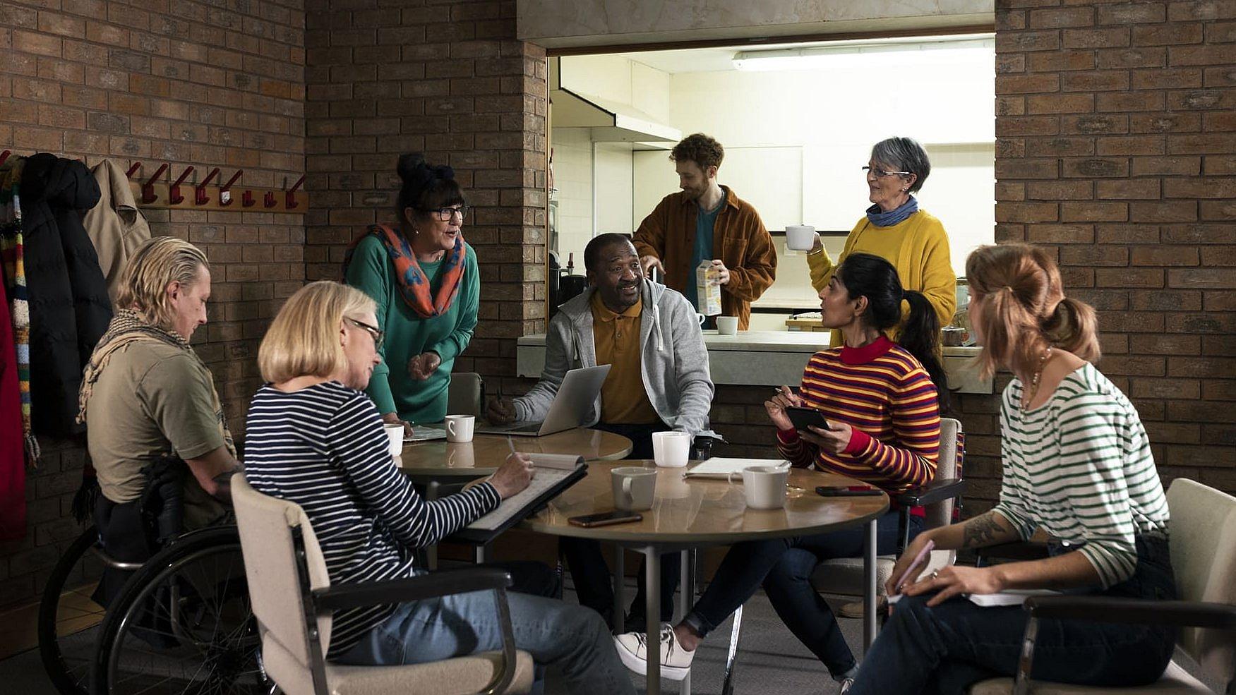 Eine Gruppe von Menschen sitzt in einem Gemeinderaum beisammen, darunter auch Menschen mit Migrationshintergrund. Sie bilden einen gemütlichen Sitzkreis.