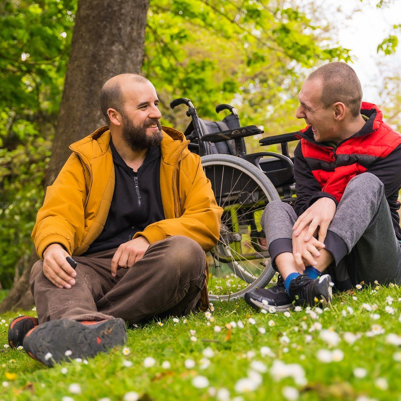 Inklusion - Seelsorge Zwei Männer sitzen im Gras, einer hat eine Behinderung. Sie unterhalten sich.