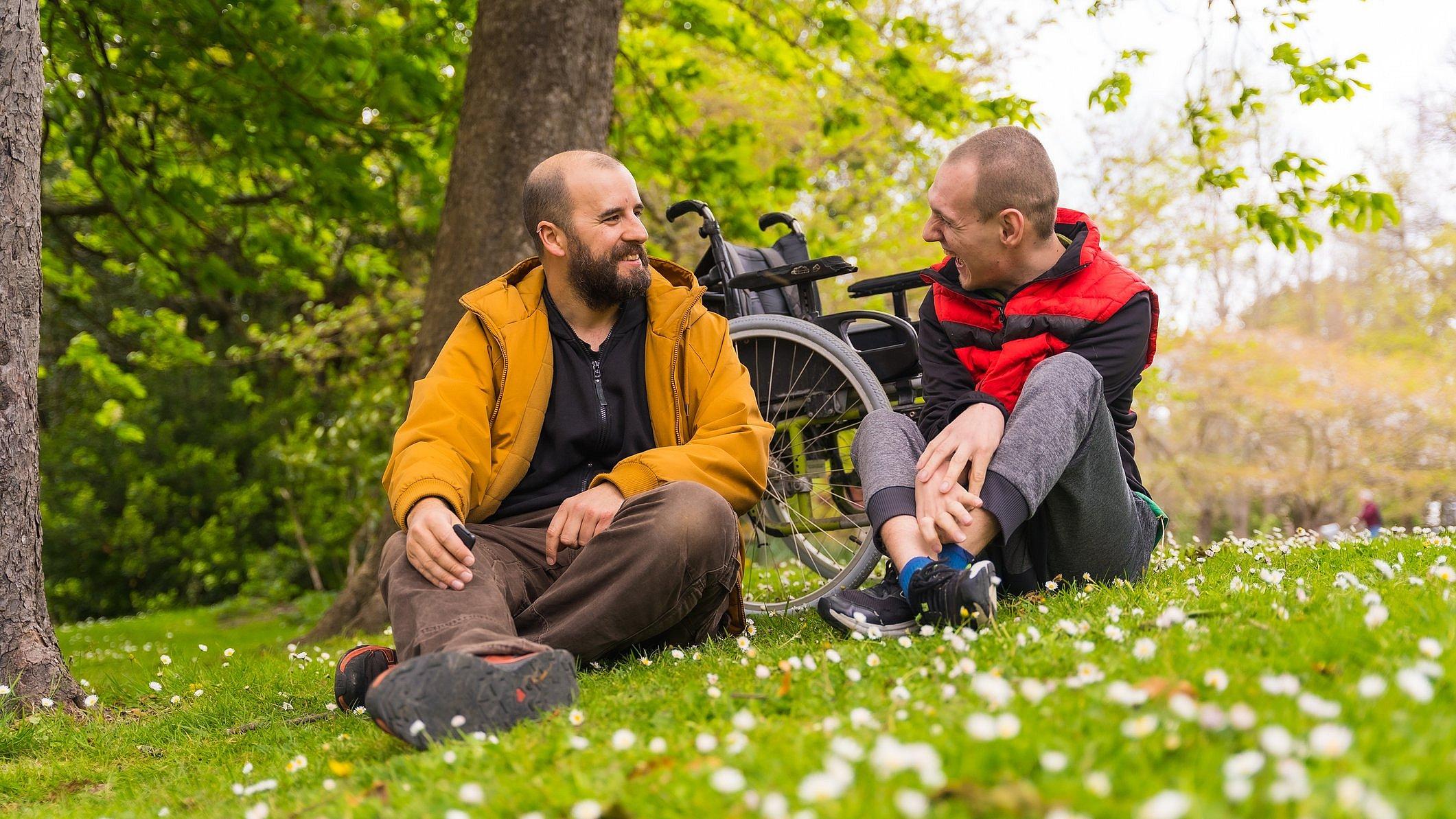 Zwei Männer sitzen im Gras, einer hat eine Behinderung. Sie unterhalten sich.