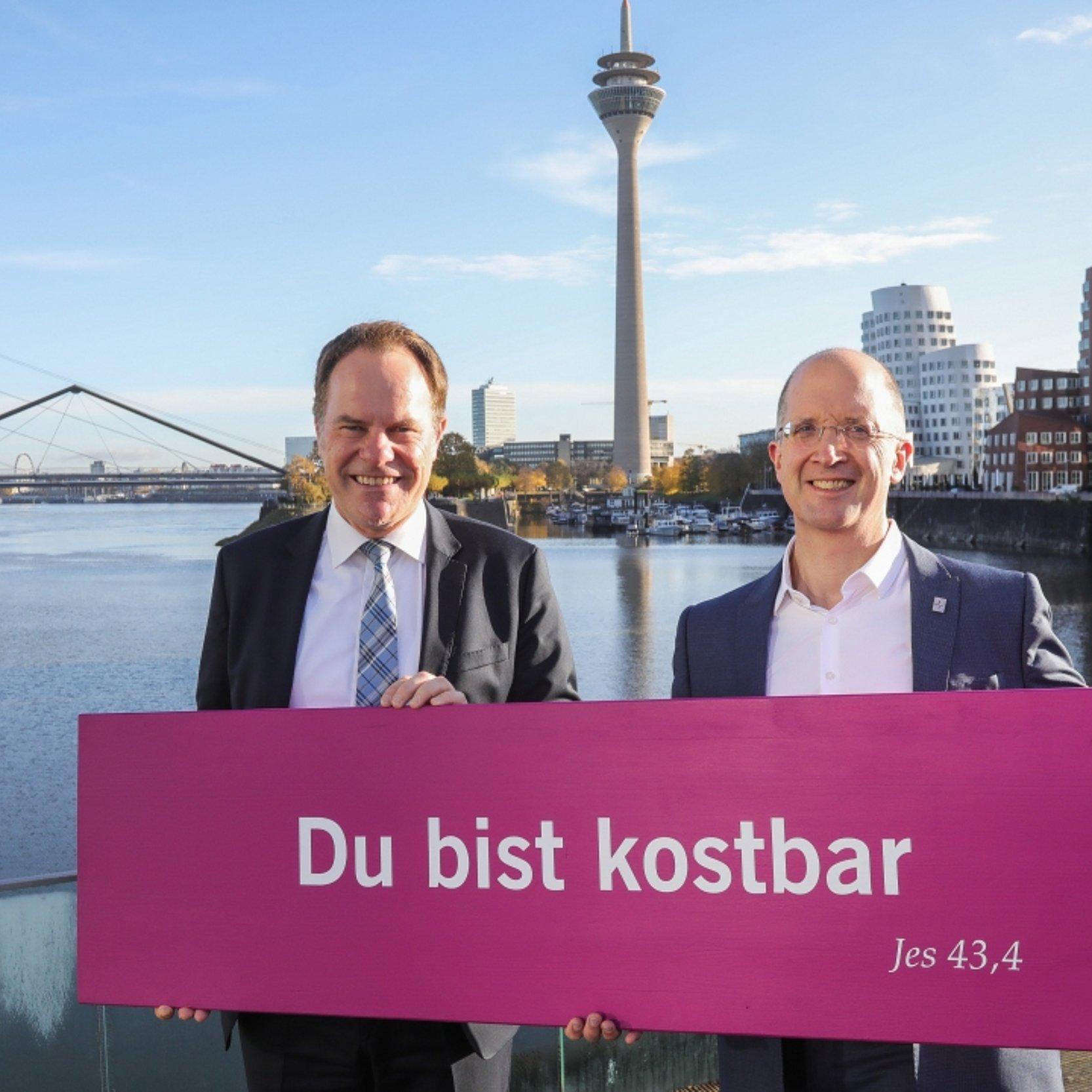 Oberbürgermeister Stephan Keller und Präses Thorsten Latzel Zwei Männer in Anzügen stehen am Rheinufer vor der Düsseldorfer Skyline mit Rheinturm im Hintergrund und halten ein pinkes Schild mit der Aufschrift „Du bist kostbar (Jes 43,4)“.