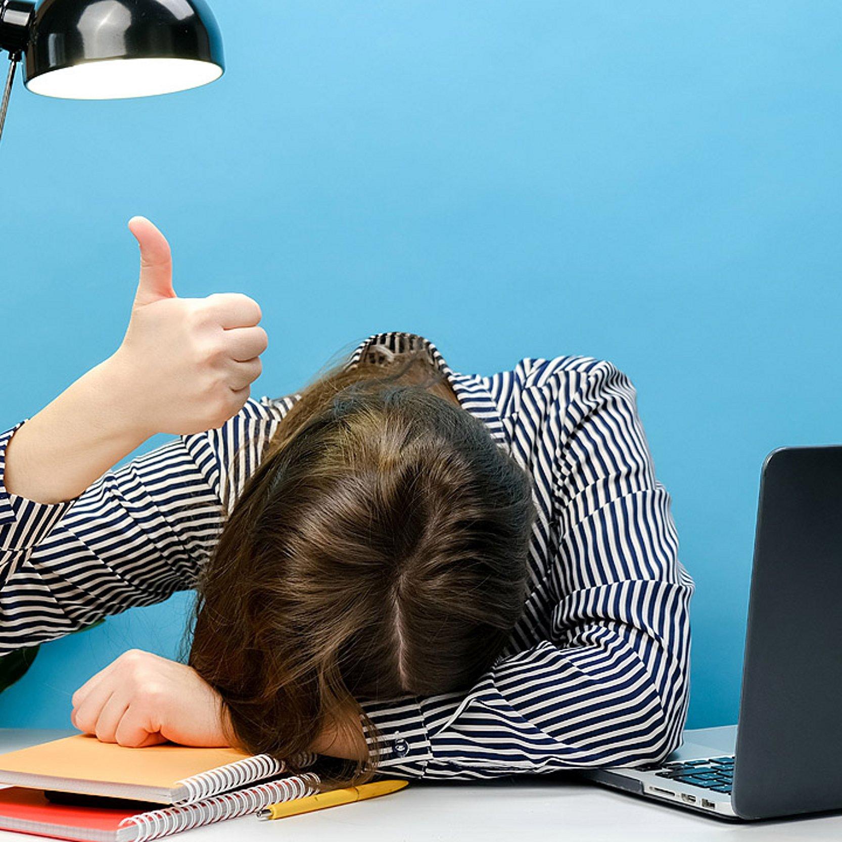 Erschöpfung Kopf eines Mannes liegt auf dem Schreibtisch, er hält aber den Daumen hoch