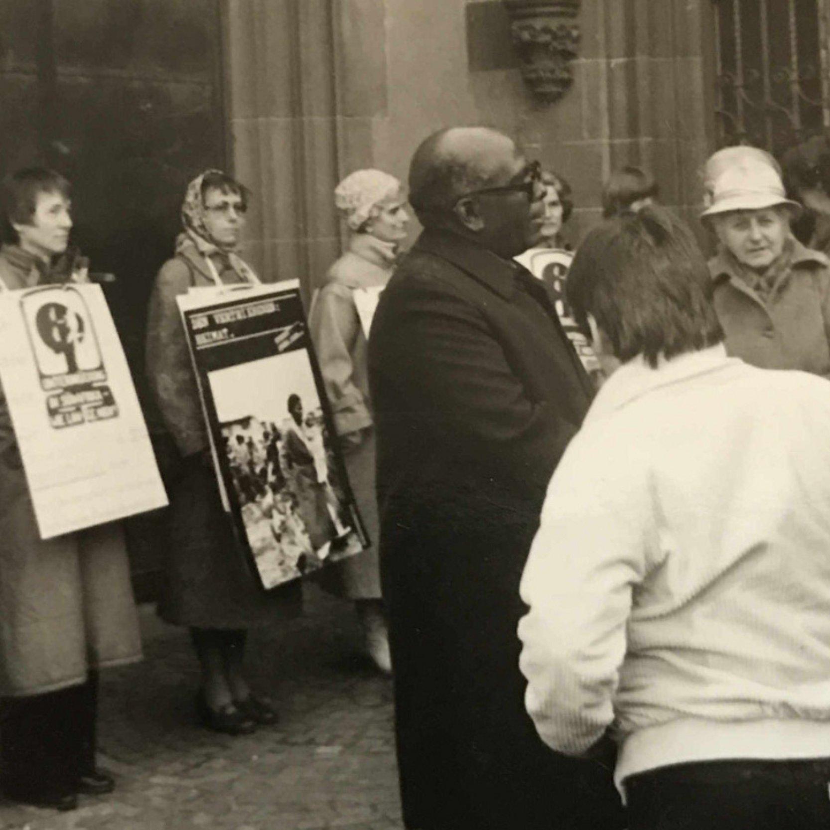 Frauen für Südafrika - Mahnwache vor dem Frankfurter Dom Frauen stehen mit Plakaten, die sie umgehängt haben vor dem Portal des Frankfurter Doms. Im Vordergrund steht eine Gruppe von Menschen und diskutert miteinander.
