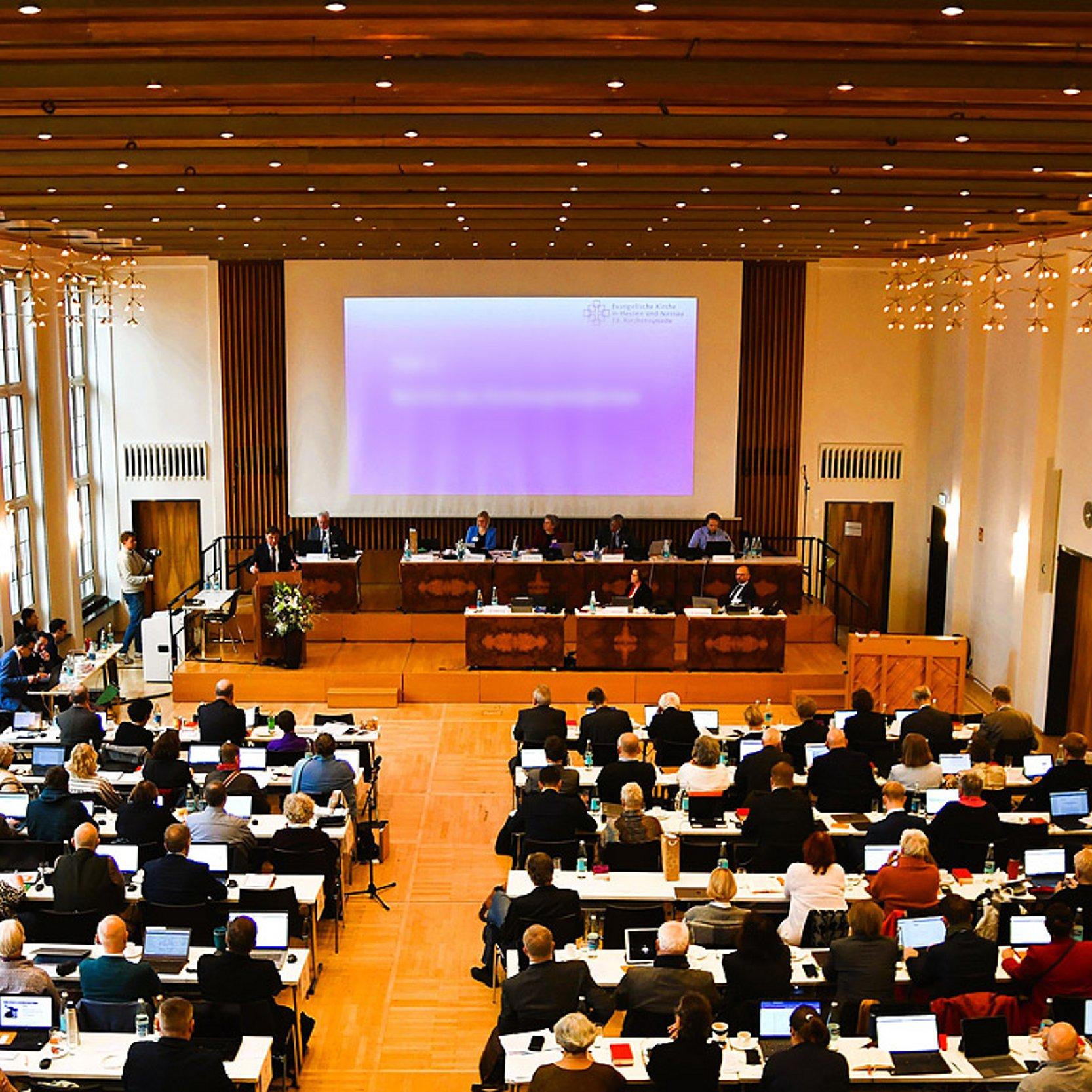 Plenum der EKHN-Synode Großer Saal mit Leinwand am Ende, davor viele Bankreihen mit Menschen