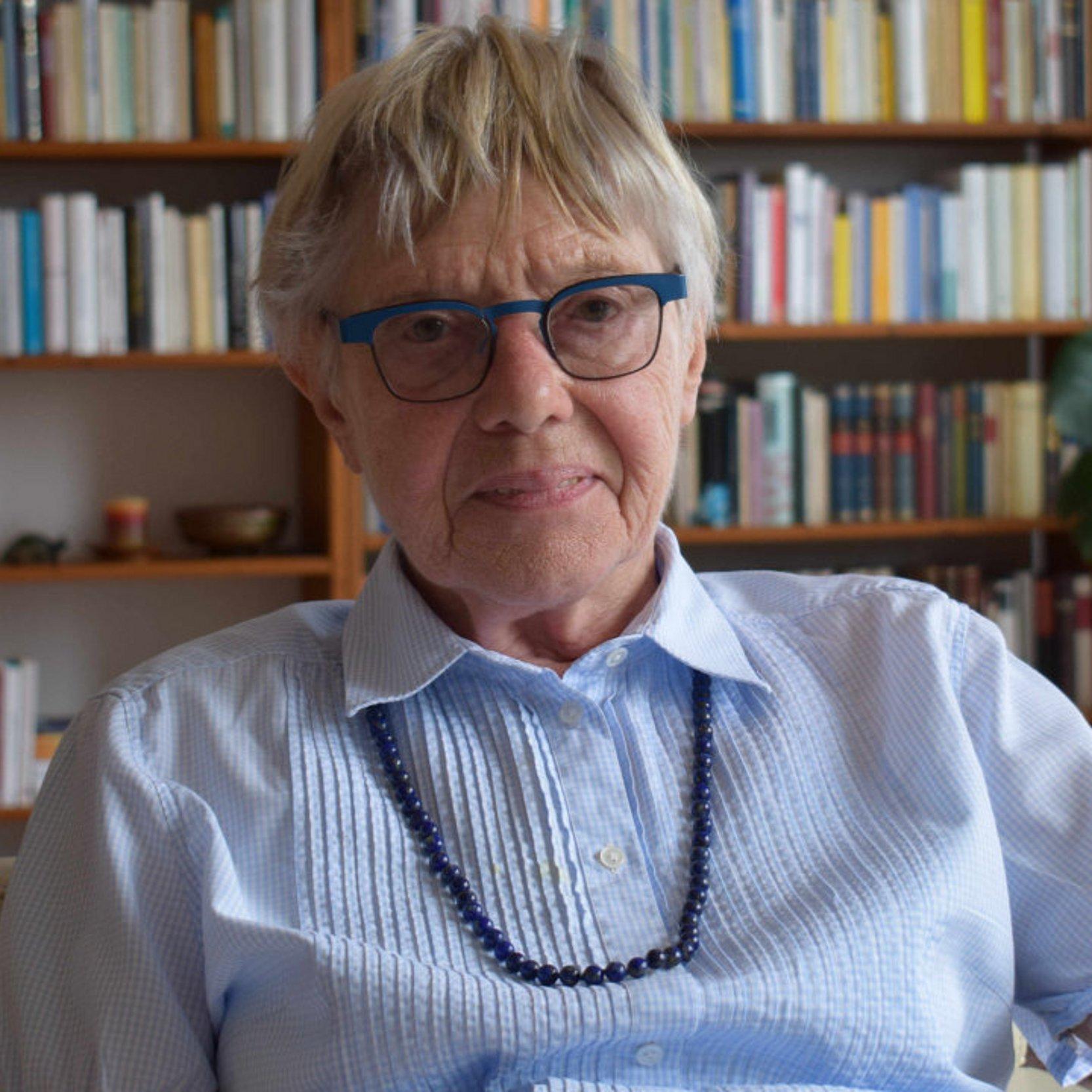 Ursula Hauptmann Ursula Hauptmann sitzt mit einer hellblauen Bluse und einer dunkelblauen Kette in ihrem Wohnzimmer, im Hintergrund ist ein Bücherregal