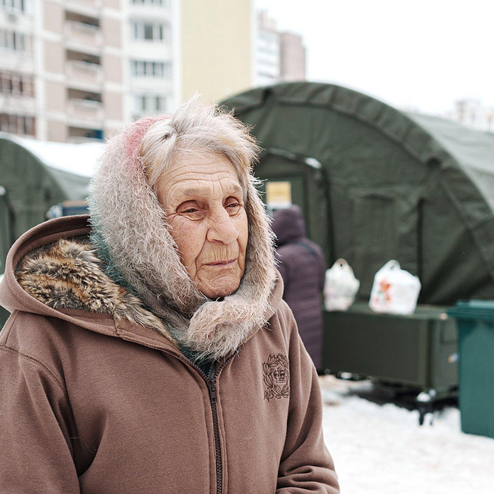 Winterhilfe Ukraine Eine ältere Dame vor einem halbrunden, langen, schwarzen Zelt, alles ist verschneit, dahinter Wohnblocks