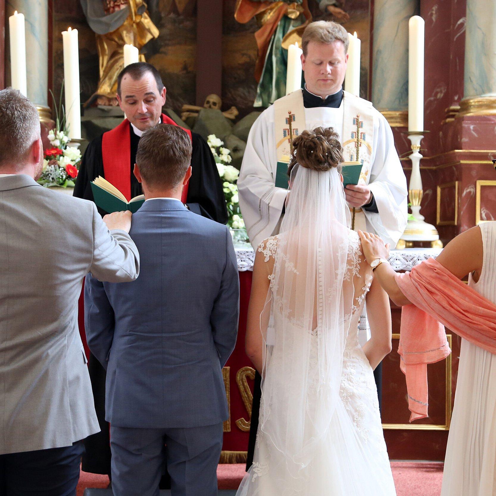 Ein Brautpaar mit Trauzeugen vor dem Altar. Vor dem Altar stehen ein katholischer und ein evangelischer Pfarrer