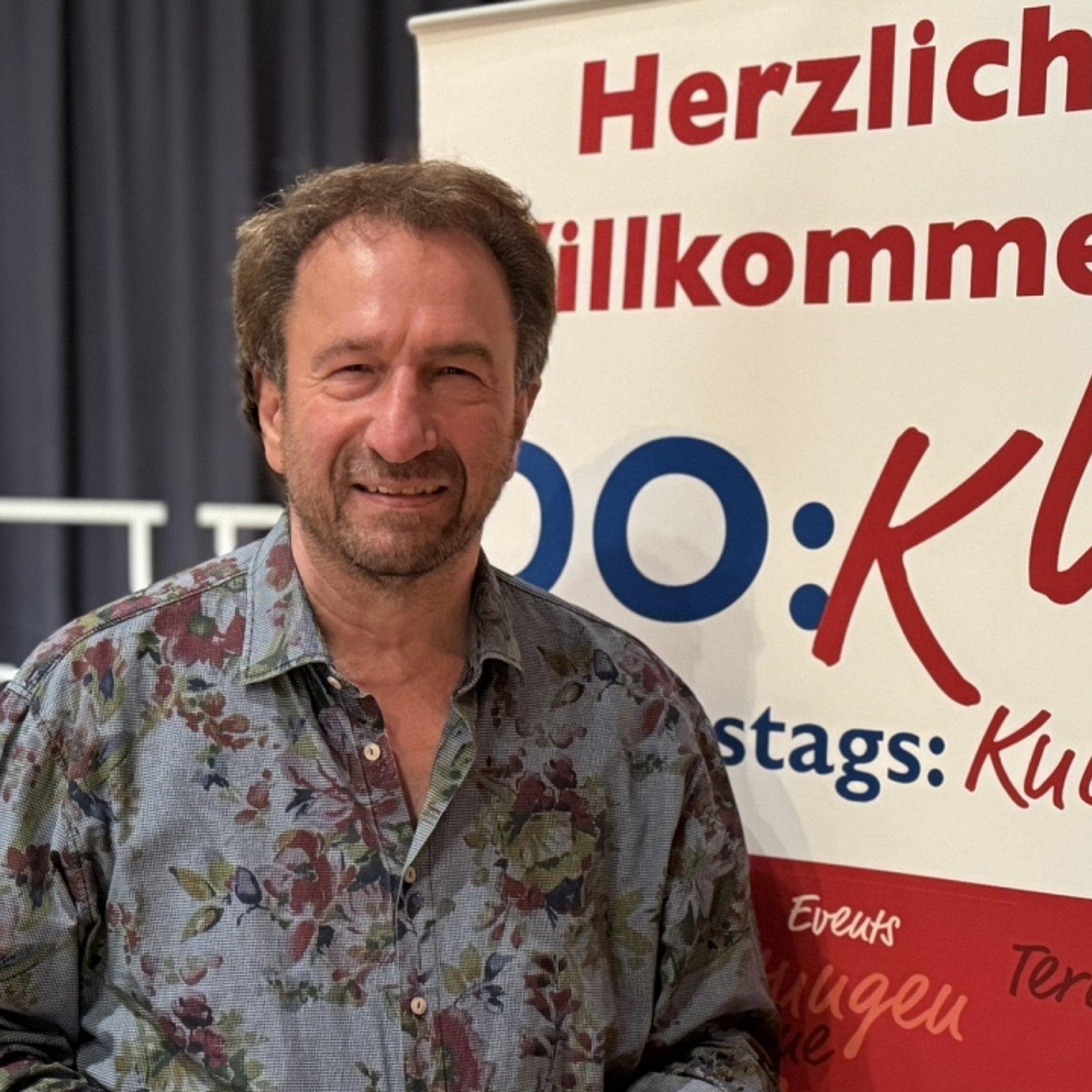 Clemens Bittlinger 2025 Portrait vor Plakat "Do:Ku" (Donnerstags:Kultur, eine Veranstaltungsreihe in Darmstadt)